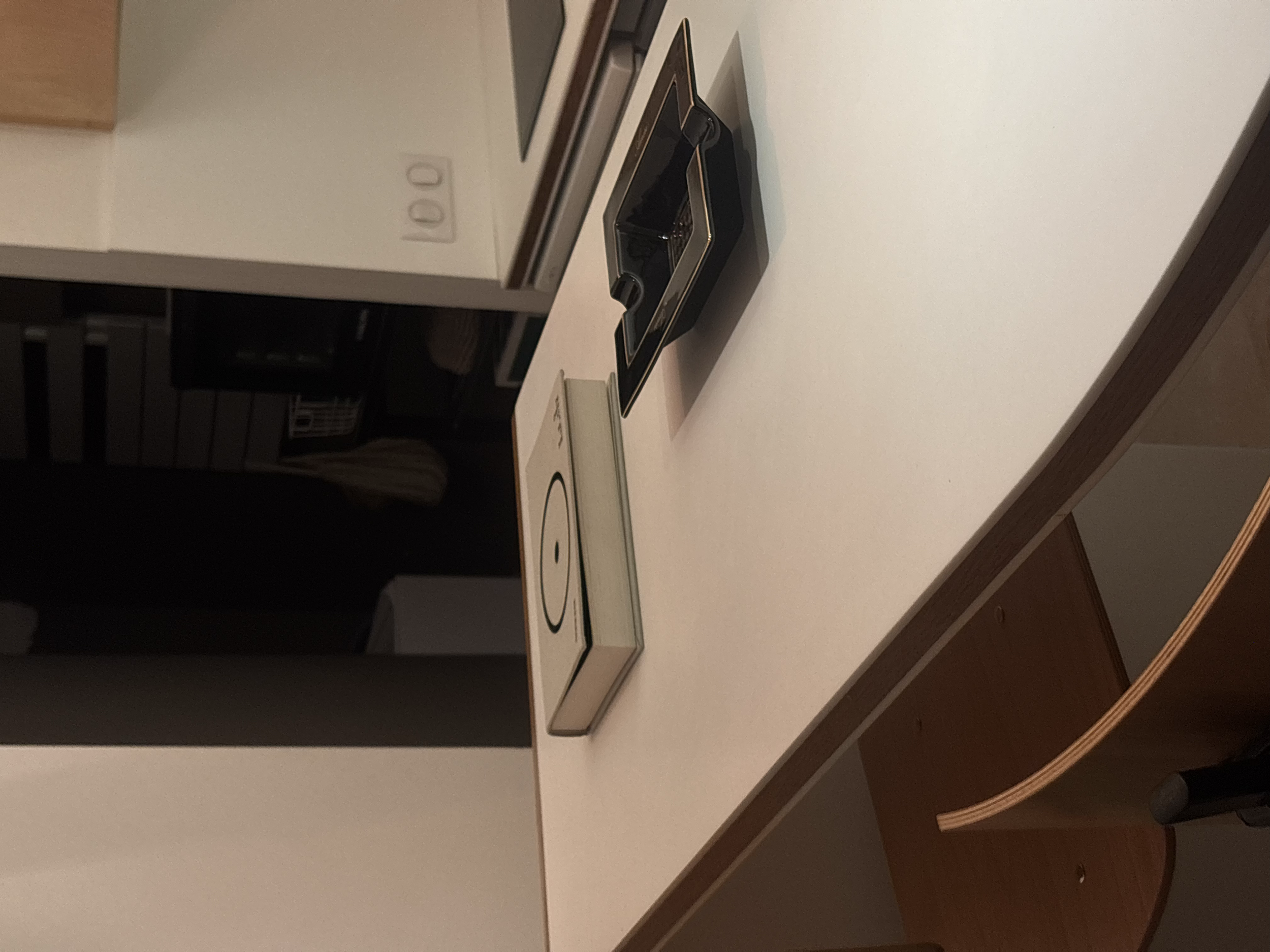 Table blanche avec une boîte de jeu de société, une tablette, et un livre. En arrière-plan, cuisine avec éléments en bois et prises électriques.