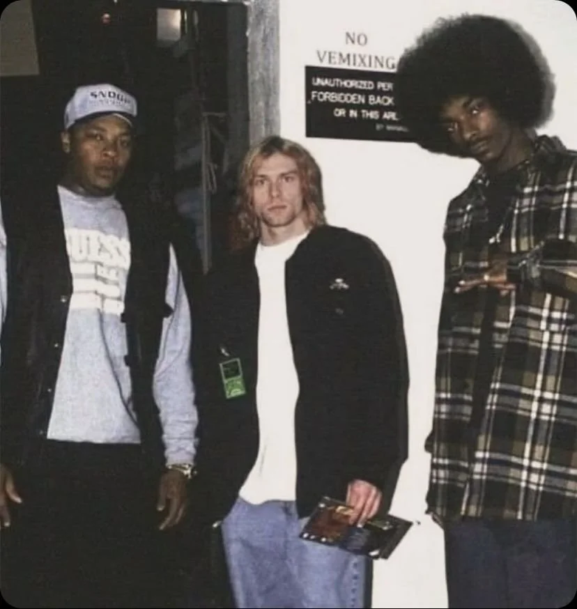 Trois hommes posent ensemble, deux d'entre eux ont de grands cheveux afro et un troisième a des cheveux longs ondulés. Ils sont dans un espace intérieur avec un panneau indiquant 'No VEMIXING' derrière eux.