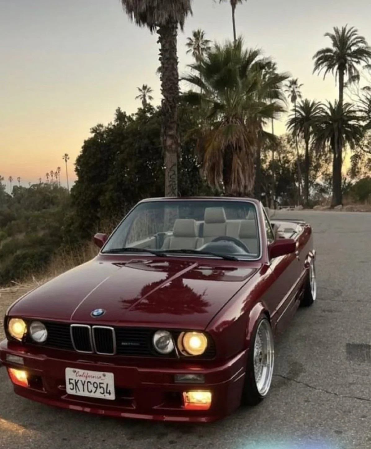Voiture BMW décapotable rouge stationnée sur une route, avec des palmiers et un ciel au coucher de soleil en arrière-plan.