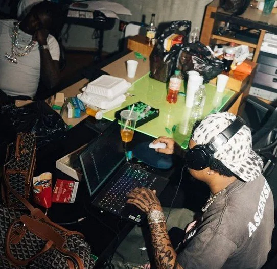 Person avec casquette et écouteurs, utilisant un ordinateur portable, assis à une table remplie de bouteilles, gobelets, nourriture à emporter et sacs, environnement intérieur