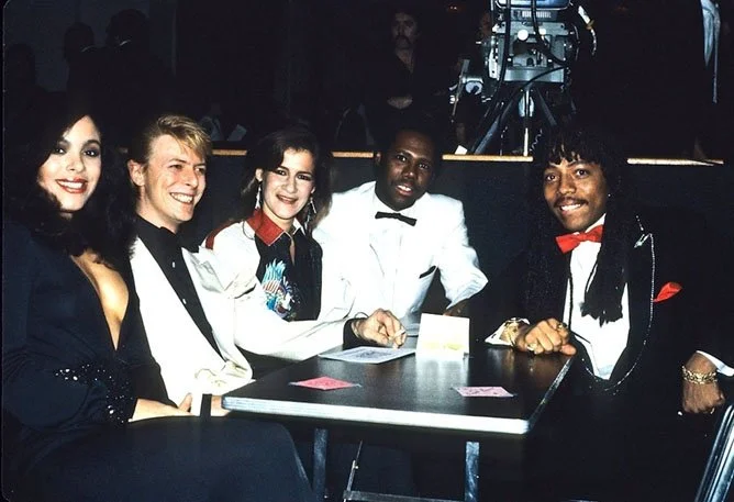 Groupe de cinq personnes assises autour d'une table lors d'un événement formel, habillées en tenues élégantes avec des costumes et des robes, sourire et posant pour la photo.