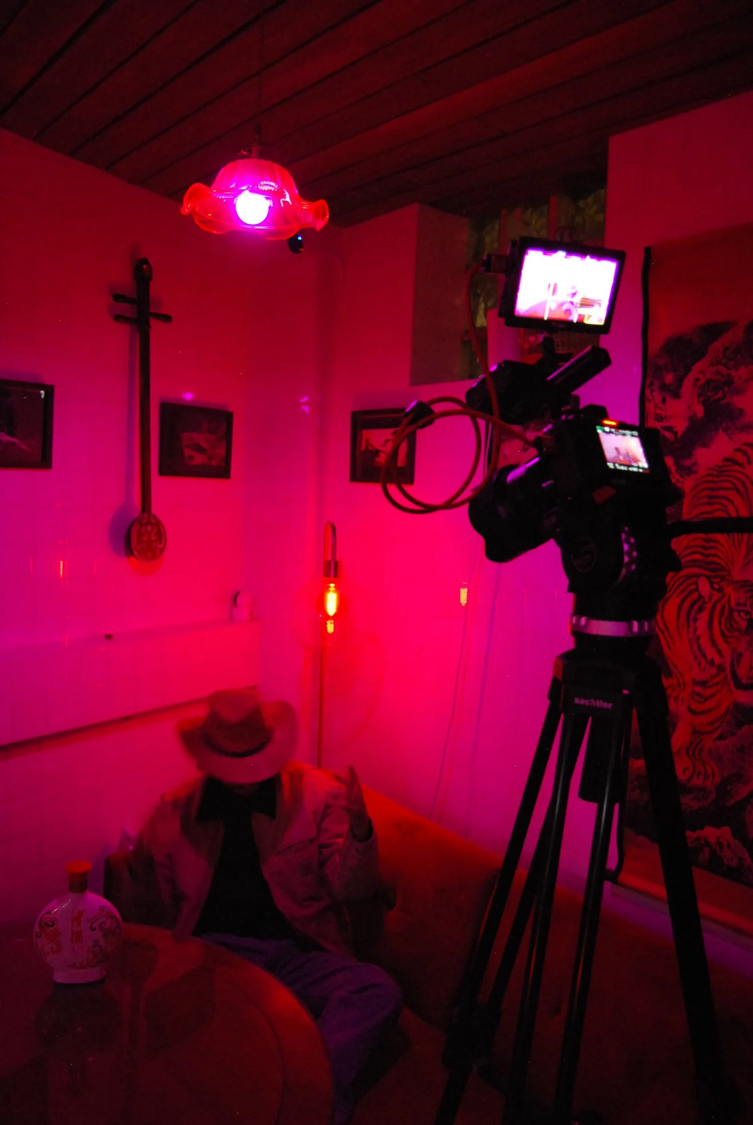 Une personne portant un chapeau et une veste claire assise sur un canapé dans une pièce éclairée en rouge. Un appareil photo ou une caméra est positionné sur un trépied, pointant vers la personne. La pièce est décorée avec des tableaux, un instrument