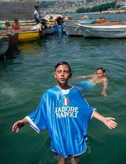 Un garçon portant un maillot de football bleu avec l'inscription "J'adore Napoli" dans une mer ou un port, avec d'autres enfants nageant et des bateaux amarrés en arrière-plan.