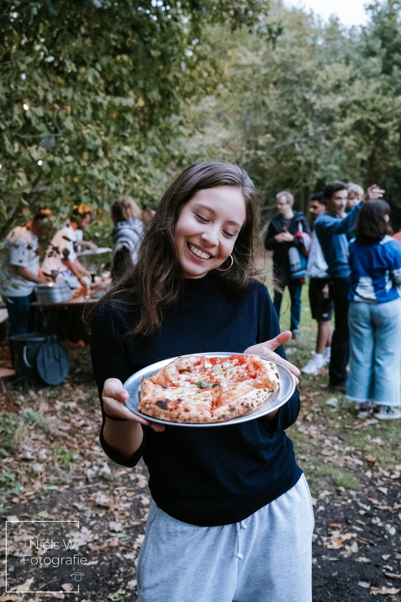 Een lachende vrouw houdt een bord met een pizza in een bosrijke omgeving, terwijl mensen op de achtergrond samen genieten.