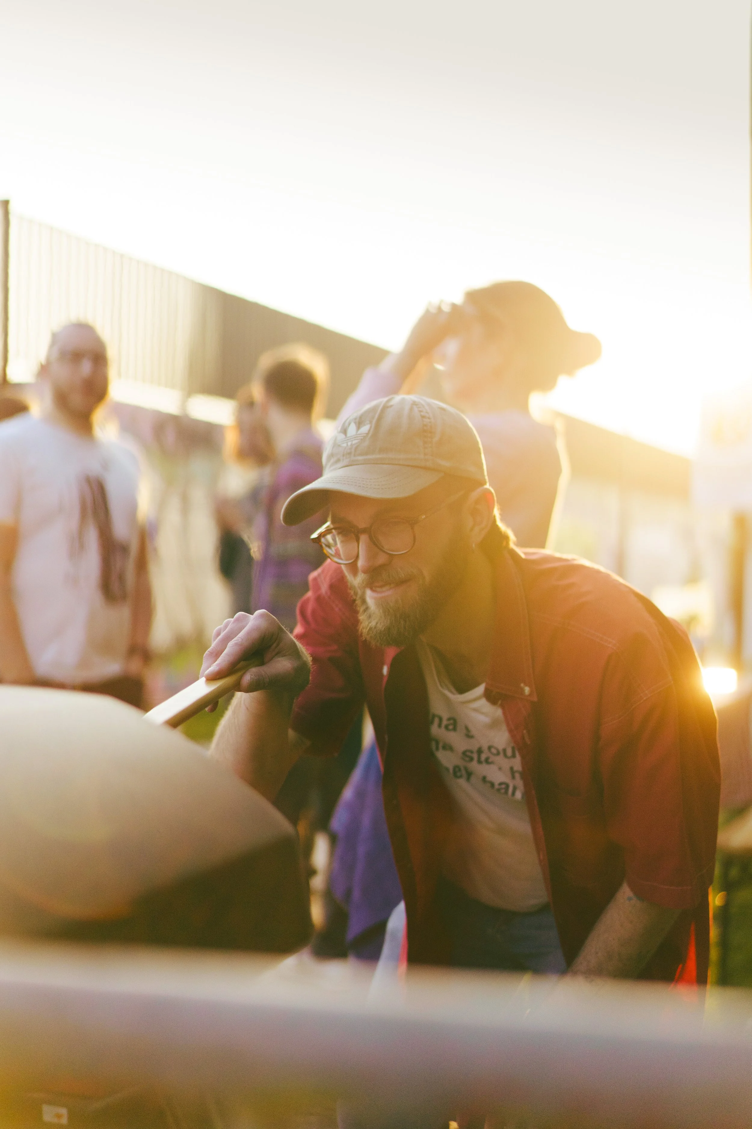 Man met bril, hoed en rood overhemd die aan een barbecue of grill roert, omgeven door andere mensen op een zomerse buitenbijeenkomst.