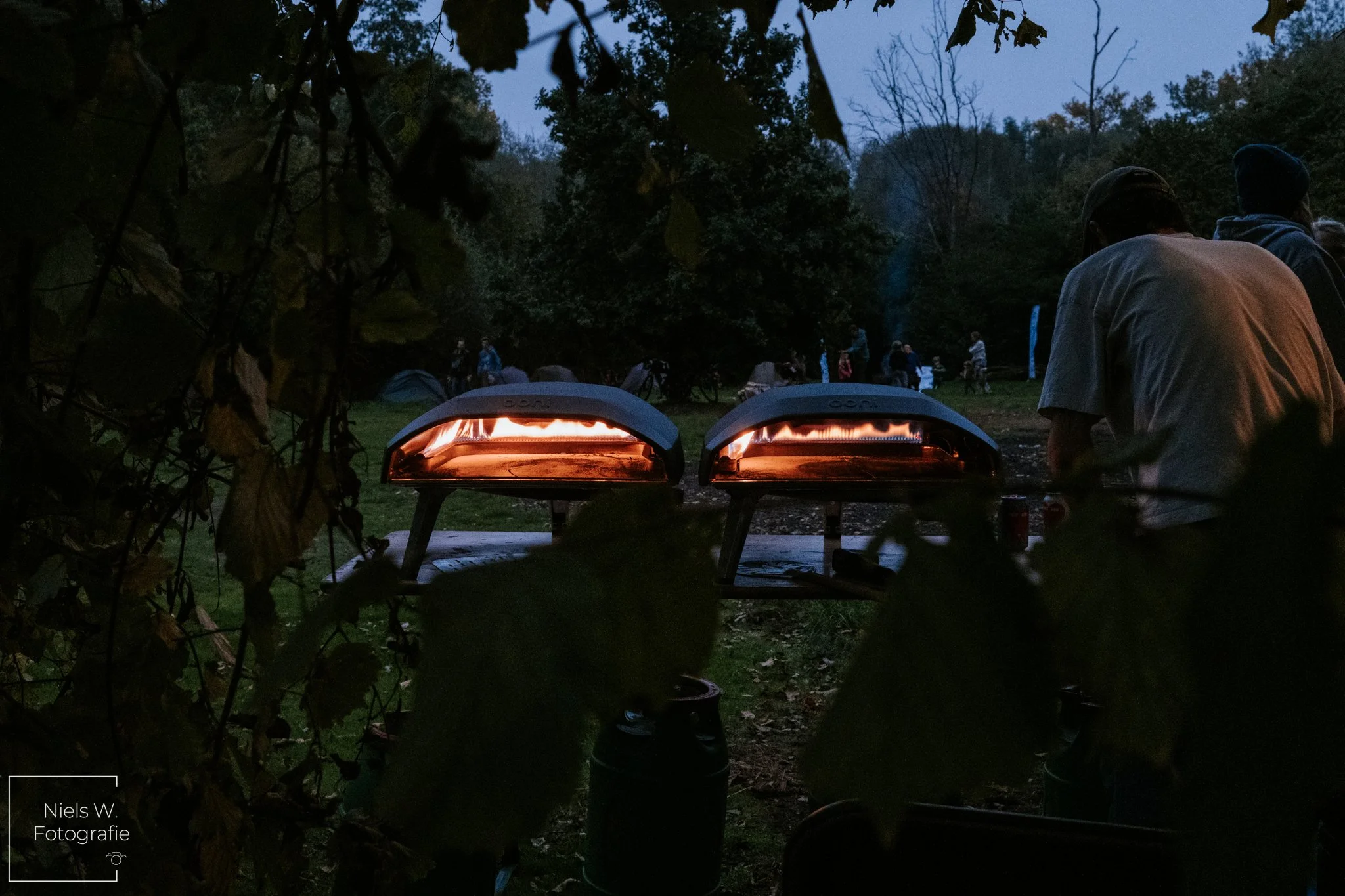 Twee kleine pizzaovens die op een tafel staan, omgeven door bomen en bladeren, met mensen op de achtergrond die zich in een buitenomgeving bevinden tijdens de schemering of avond.