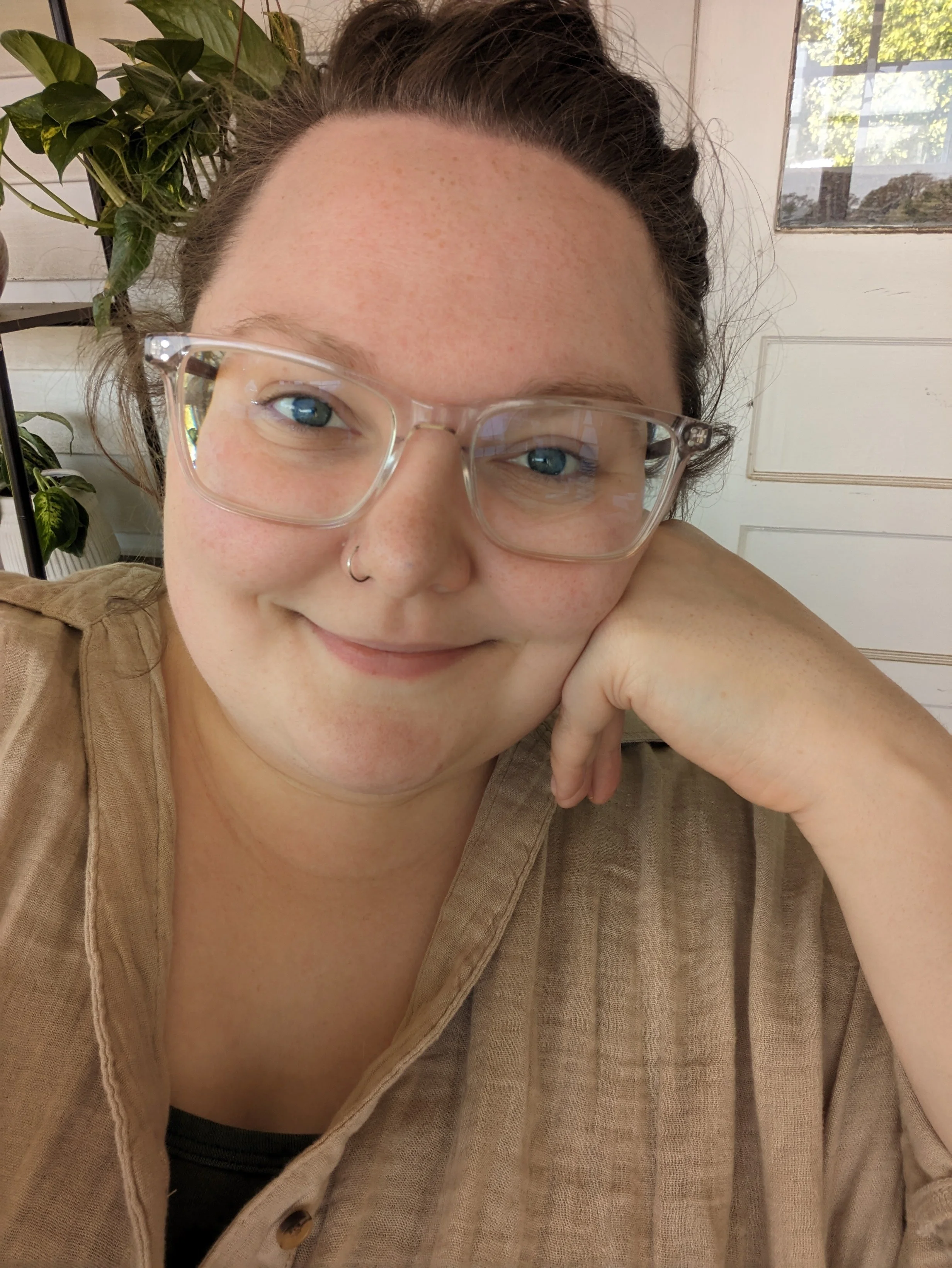 A woman with curly hair and glasses, smiling, resting her chin on her hand, in a room with a window and a plant.