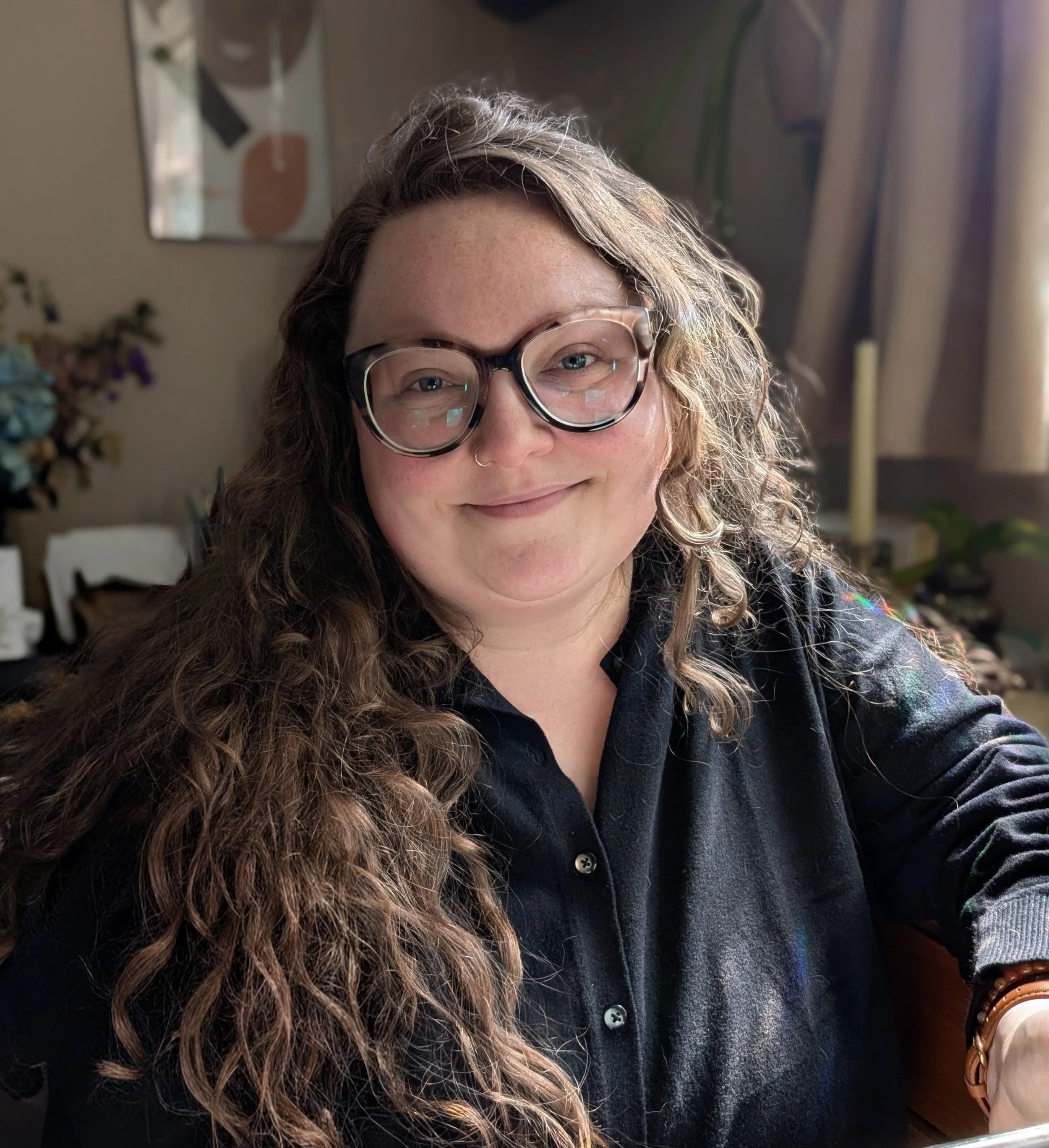 Close-up of a woman with long, curly hair wearing glasses with tortoise shell frames, smiling while sitting indoors near a window with sunlight, a mirror, and a blurred background of houseplants and furniture.