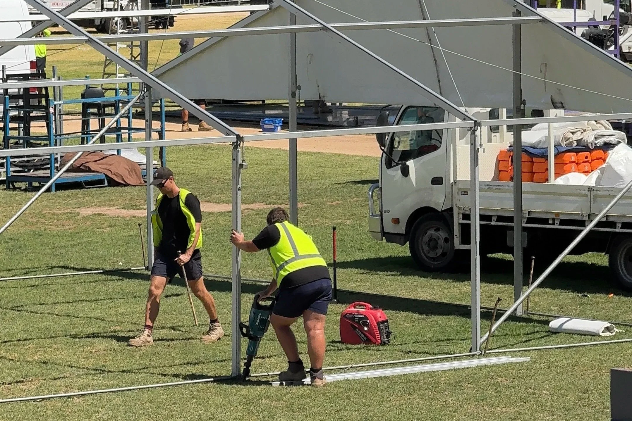 Two construction workers setting up a metal frame for a tent or structure in an outdoor event space. One worker is using a power tool, while the other is walking with a cane. There is a white truck and various equipment in the background.
