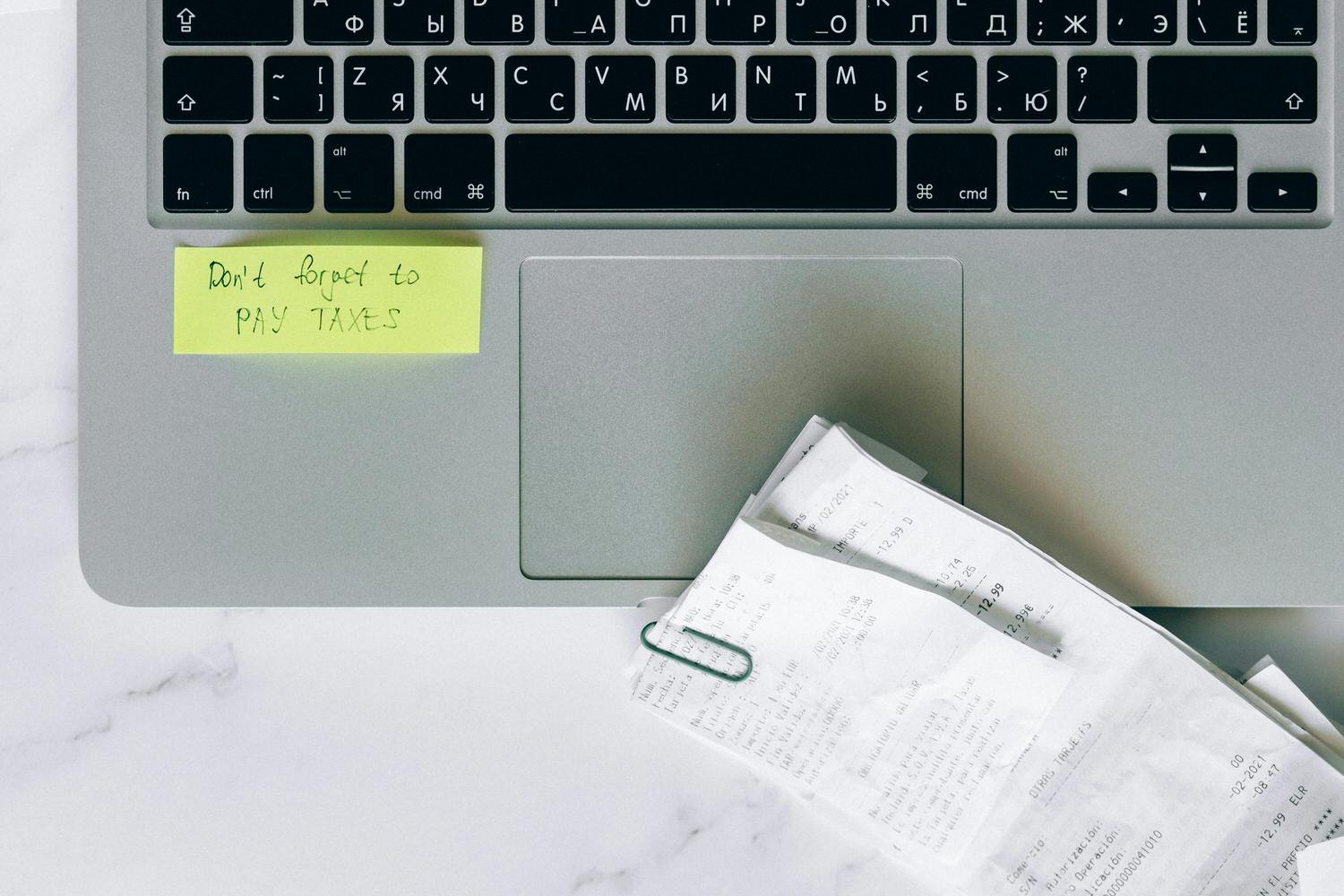 Laptop keyboard with a yellow sticky note that says 'Don't forget to PAY TAXES' and a crumpled receipt paper held with a paperclip on a white surface.