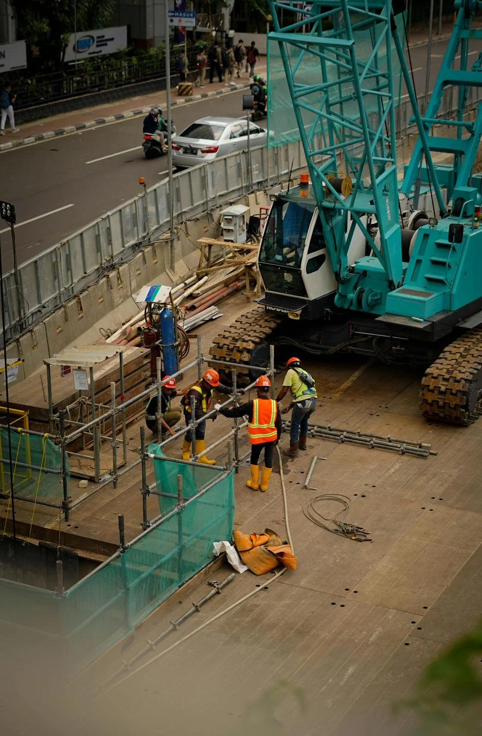 Construction workers in safety gear working near a large crane on a construction site, with a busy street and scooter traffic in the background.