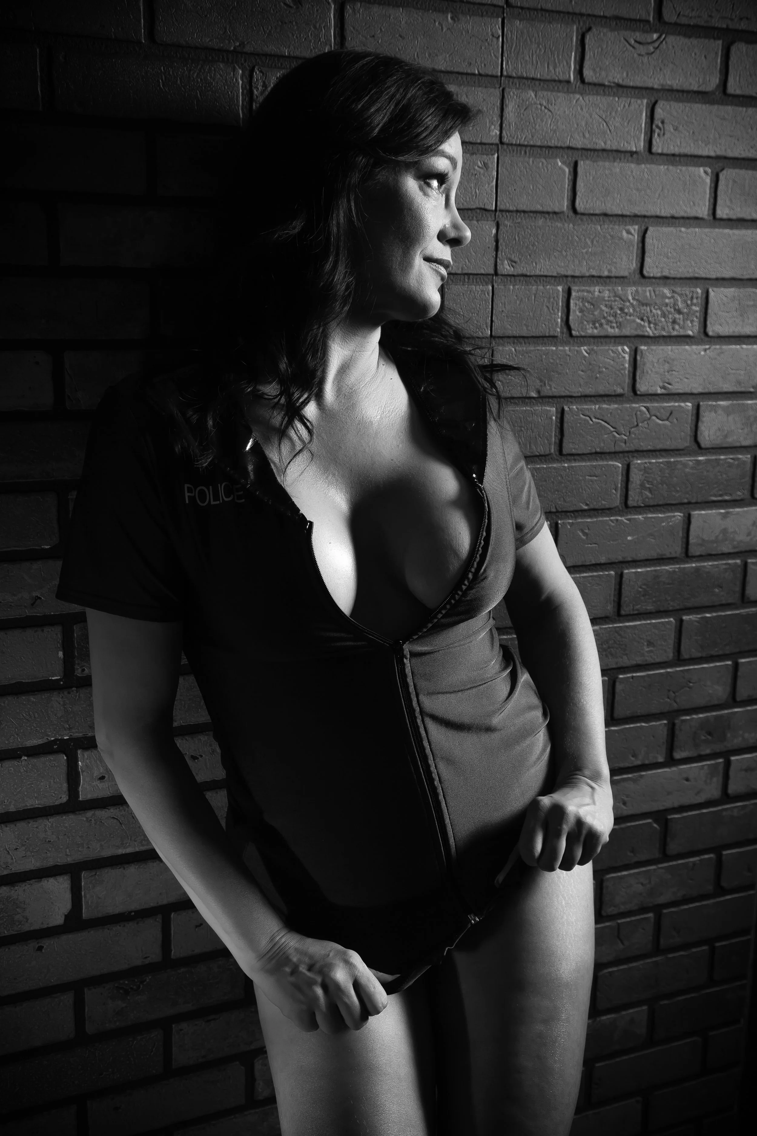 Black and white photo of a woman in a police uniform front of brick wall, looking to the side, with hair down, highlighting facial features and cleavage.