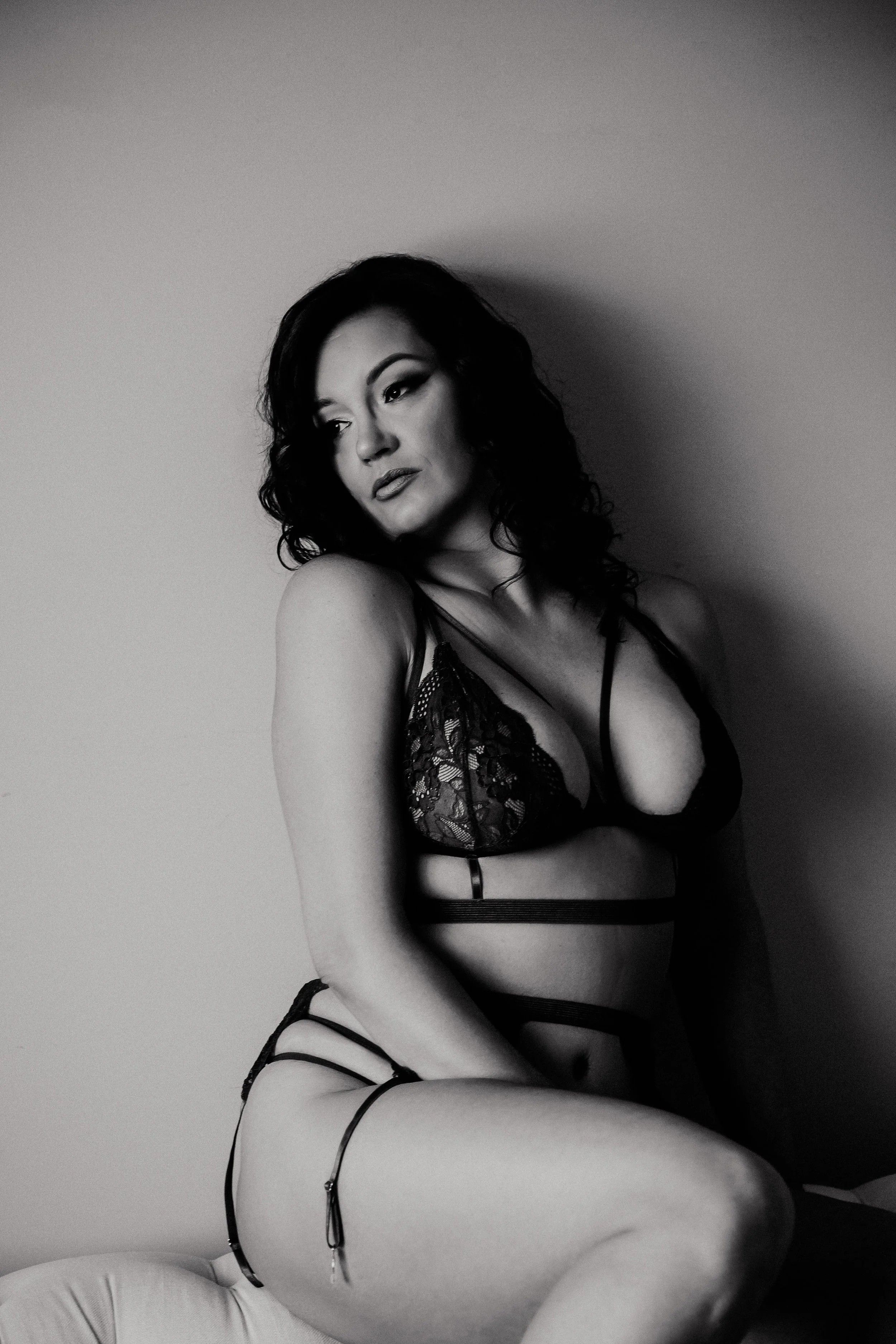 Black and white photo of a woman with dark curly hair wearing a revealing lace lingerie outfit, seated on a bed.