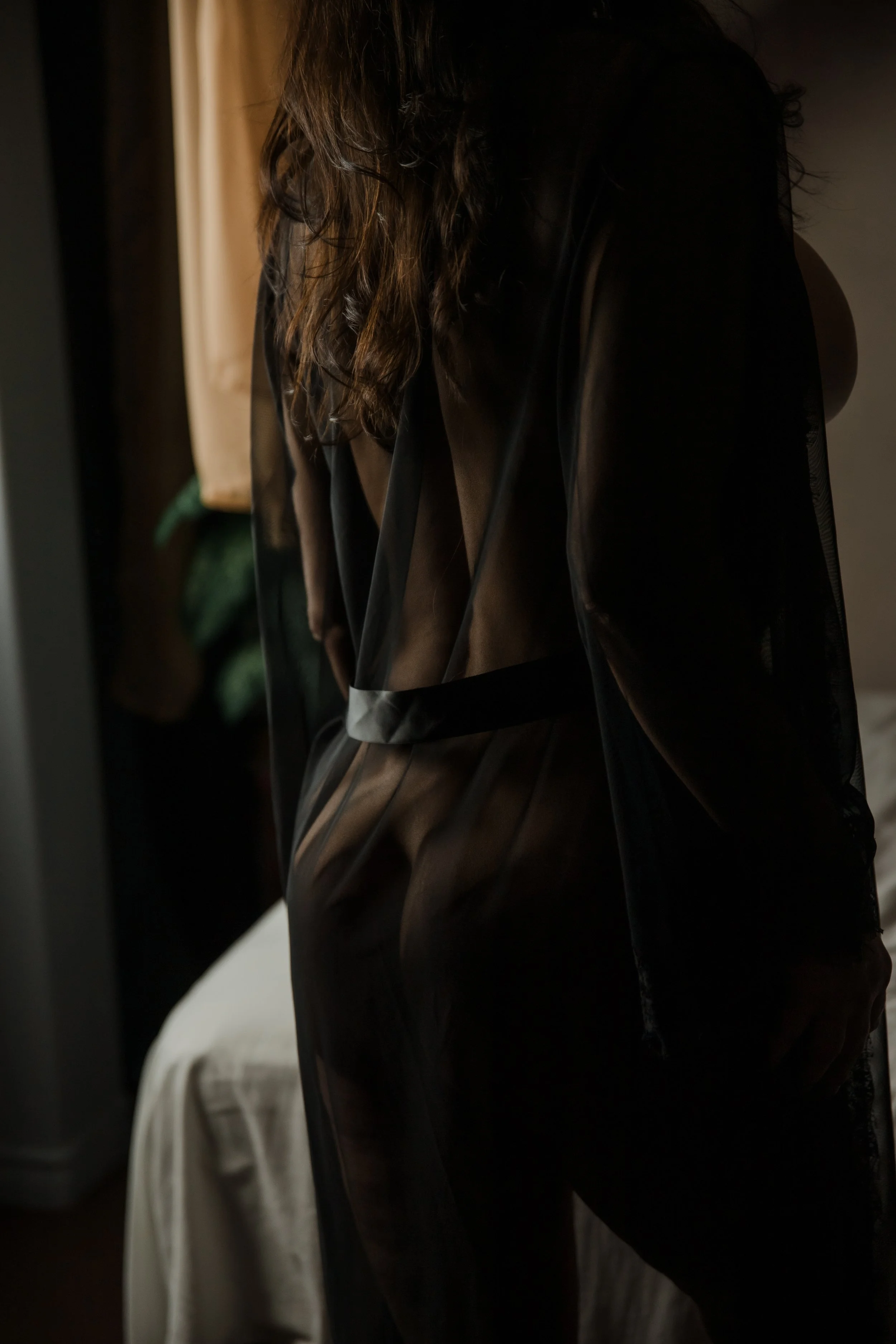 A woman with long hair wearing a sheer black robe standing indoors near a mirror with natural light.
