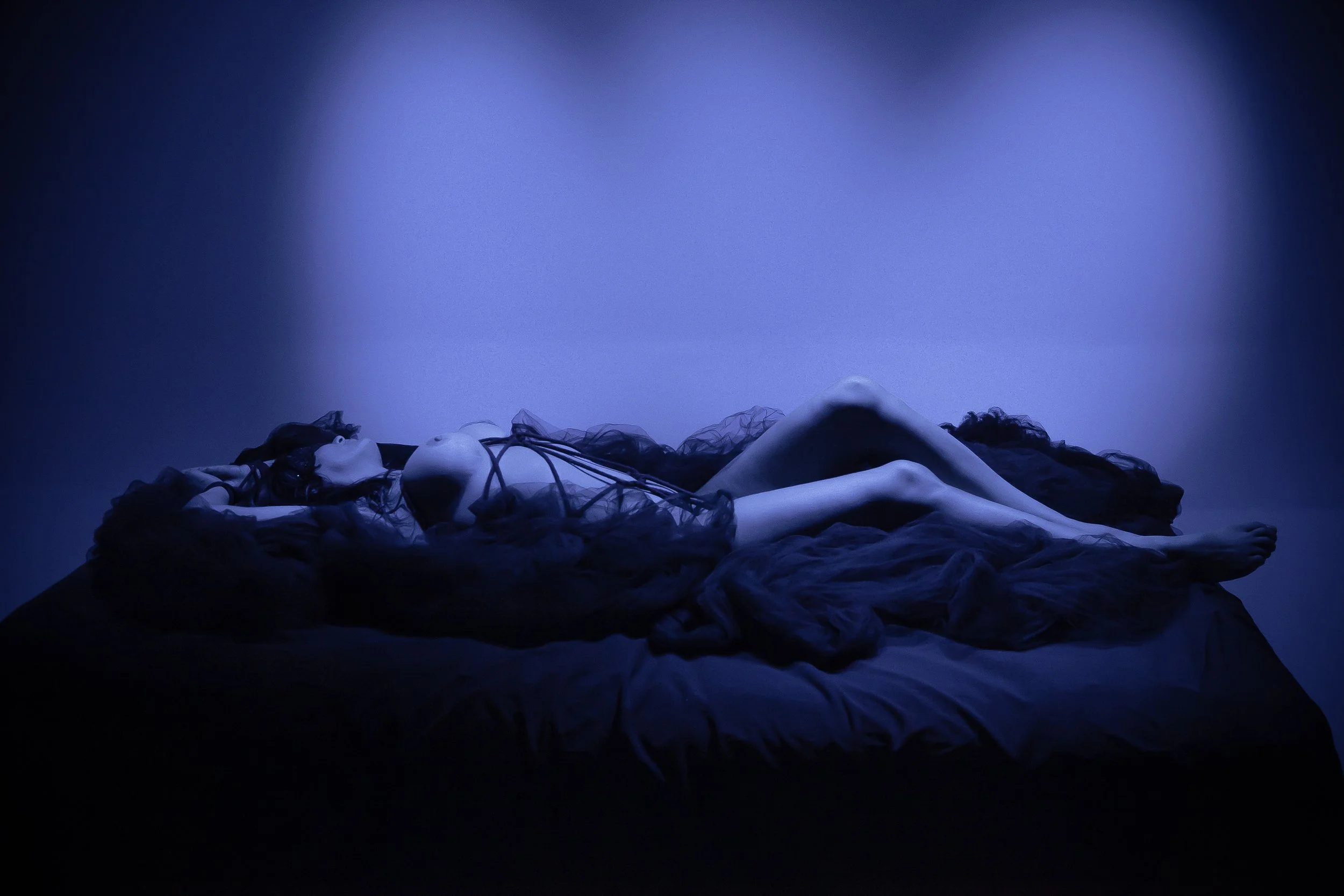 A woman lying on a bed covered in dark fabric, illuminated by blue light with a plain, slightly gradient background.