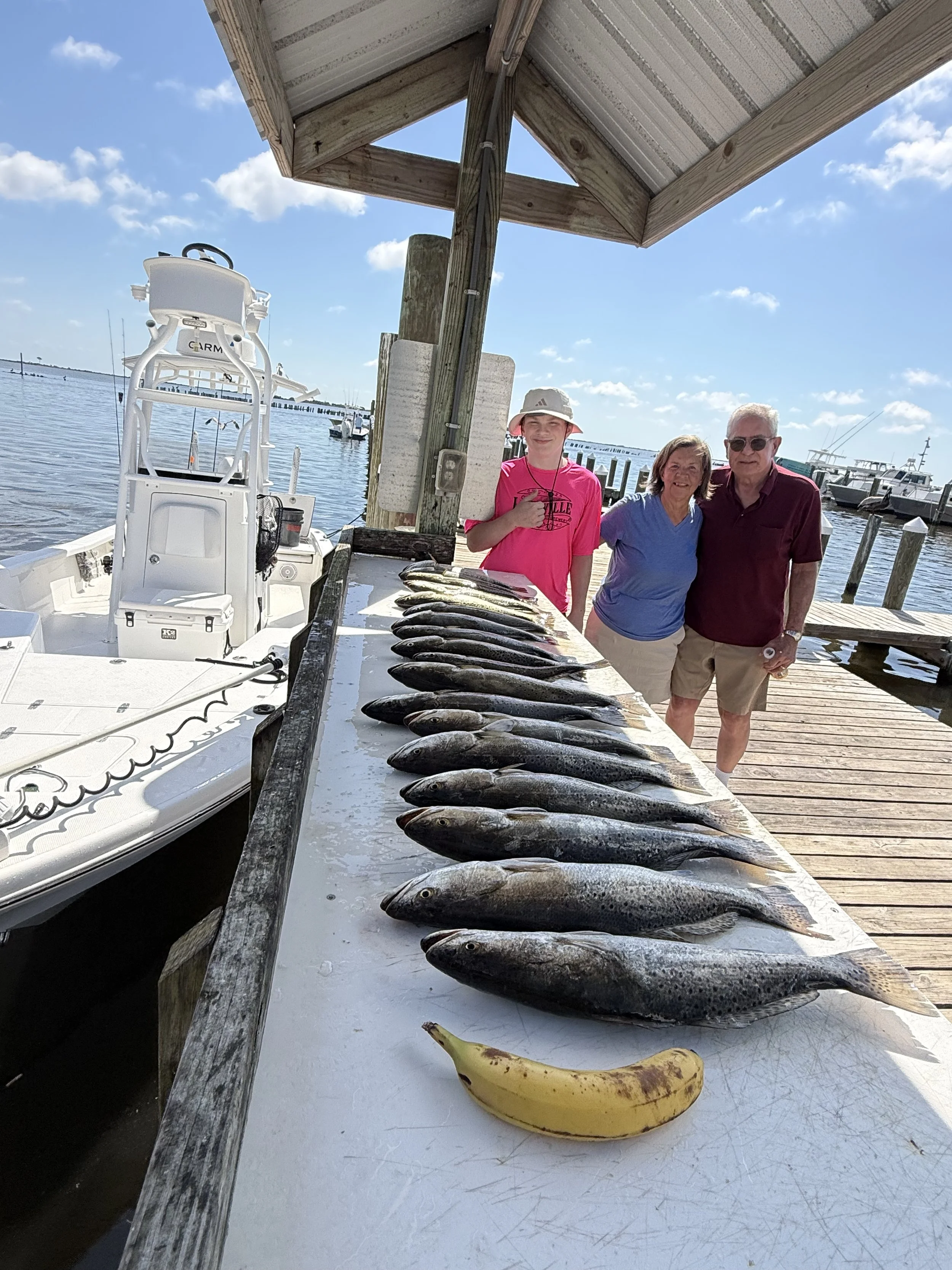 A table of caught fish, a banana, and a boat at a marina with a wooden dock and three people posing in front of the fish, under a canopy on a bright day.
