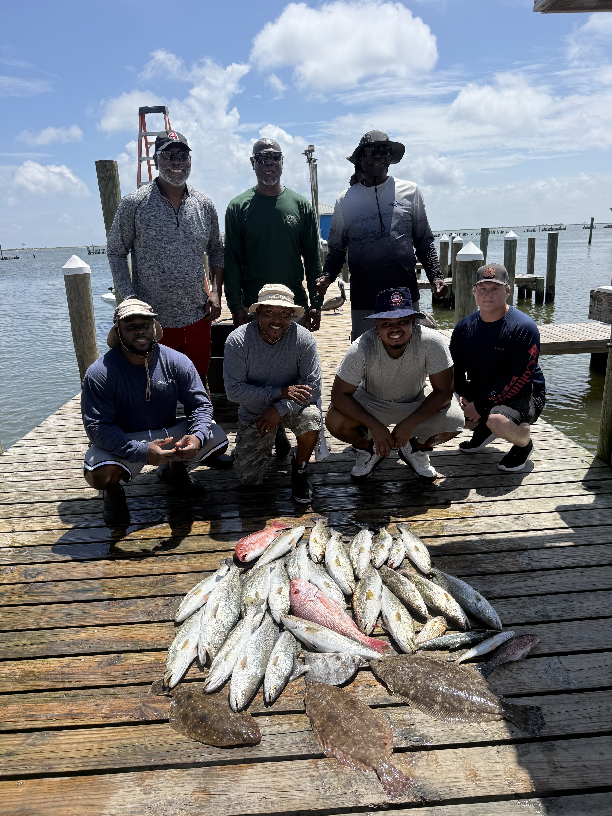 Group of seven men posing on a wooden dock by the water with caught fish laid out in front of them, including various species like red snapper and flounder, under a partly cloudy sky.