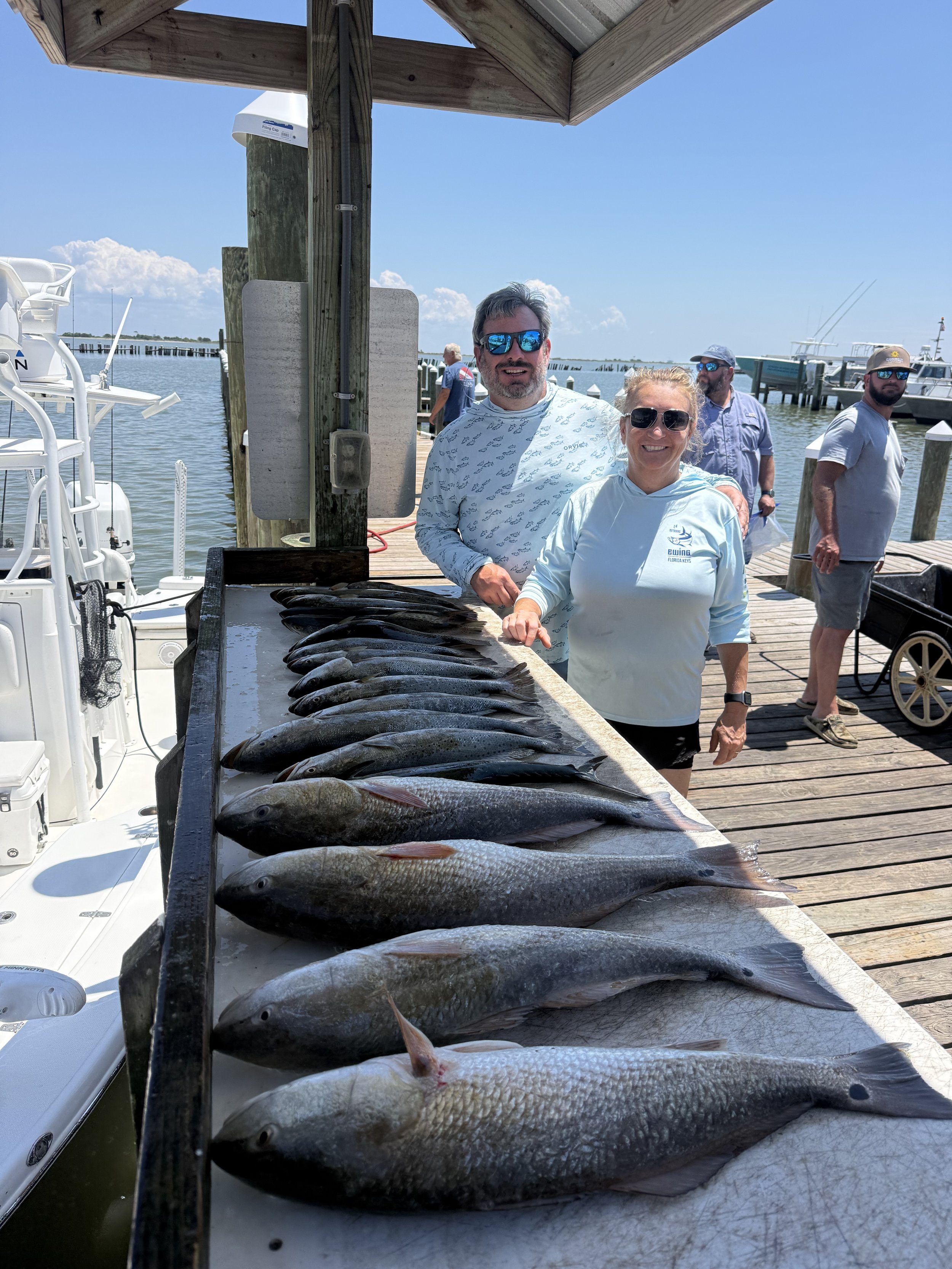 A group of people at a dock with freshly caught fish laid out on a table. The scene includes boats and a blue sky with some clouds in the background.