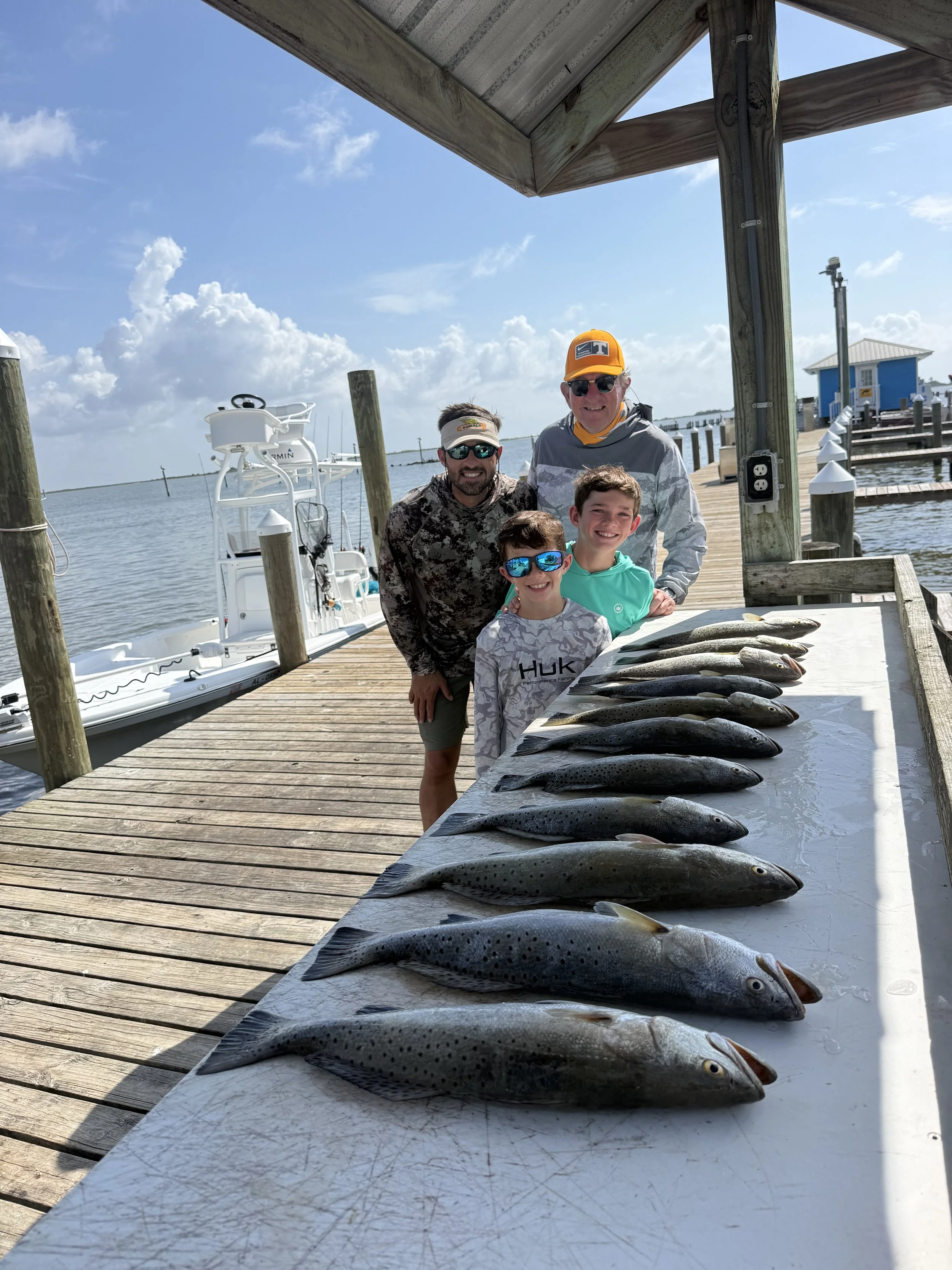 Four fish laid out on a white table on a dock, with four people smiling behind it, and boats docked nearby on a sunny day.
