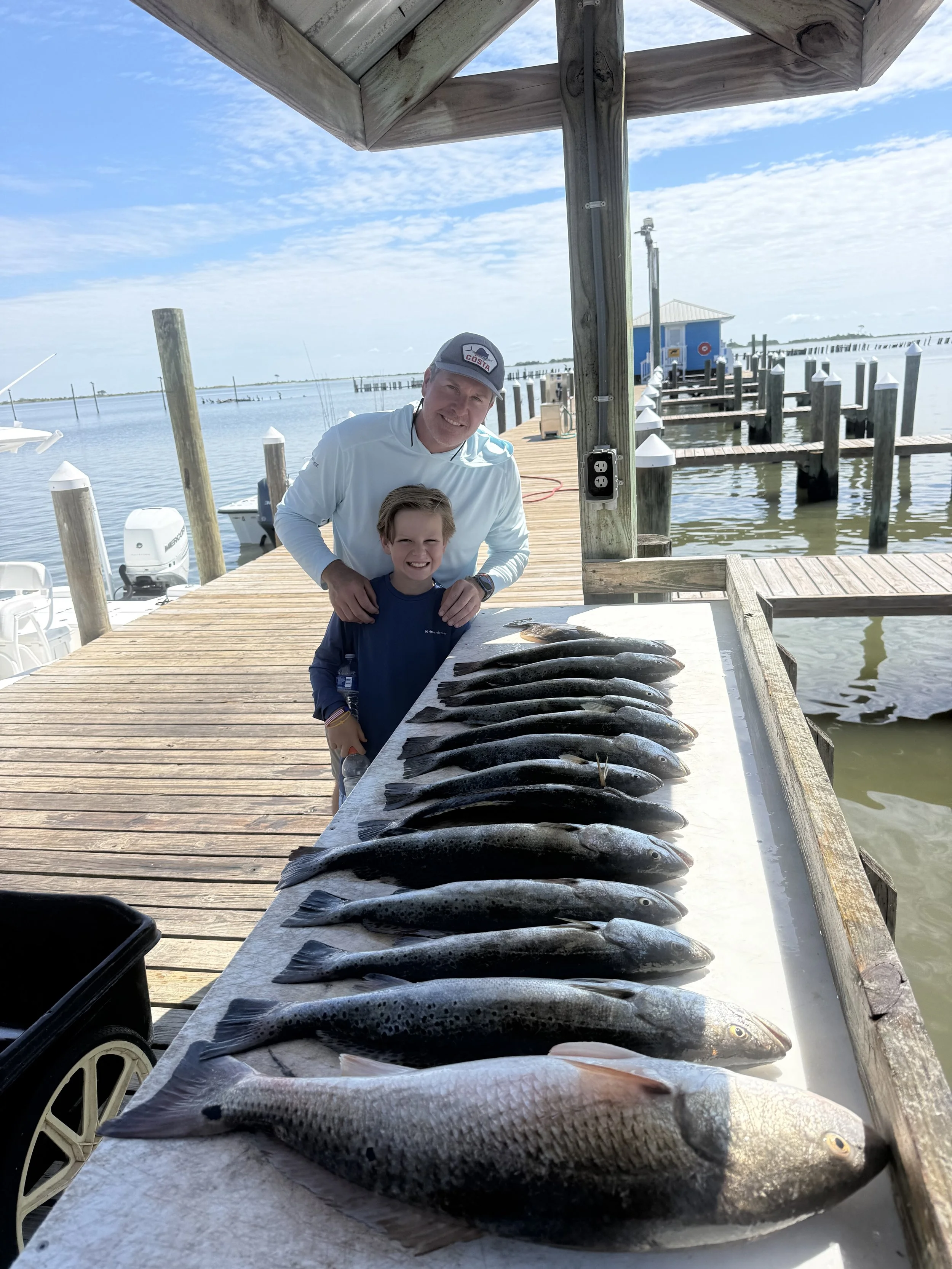 A man and a young boy smiling at a dock next to a row of freshly caught fish laid out on a white surface, with a lake and boats in the background.
