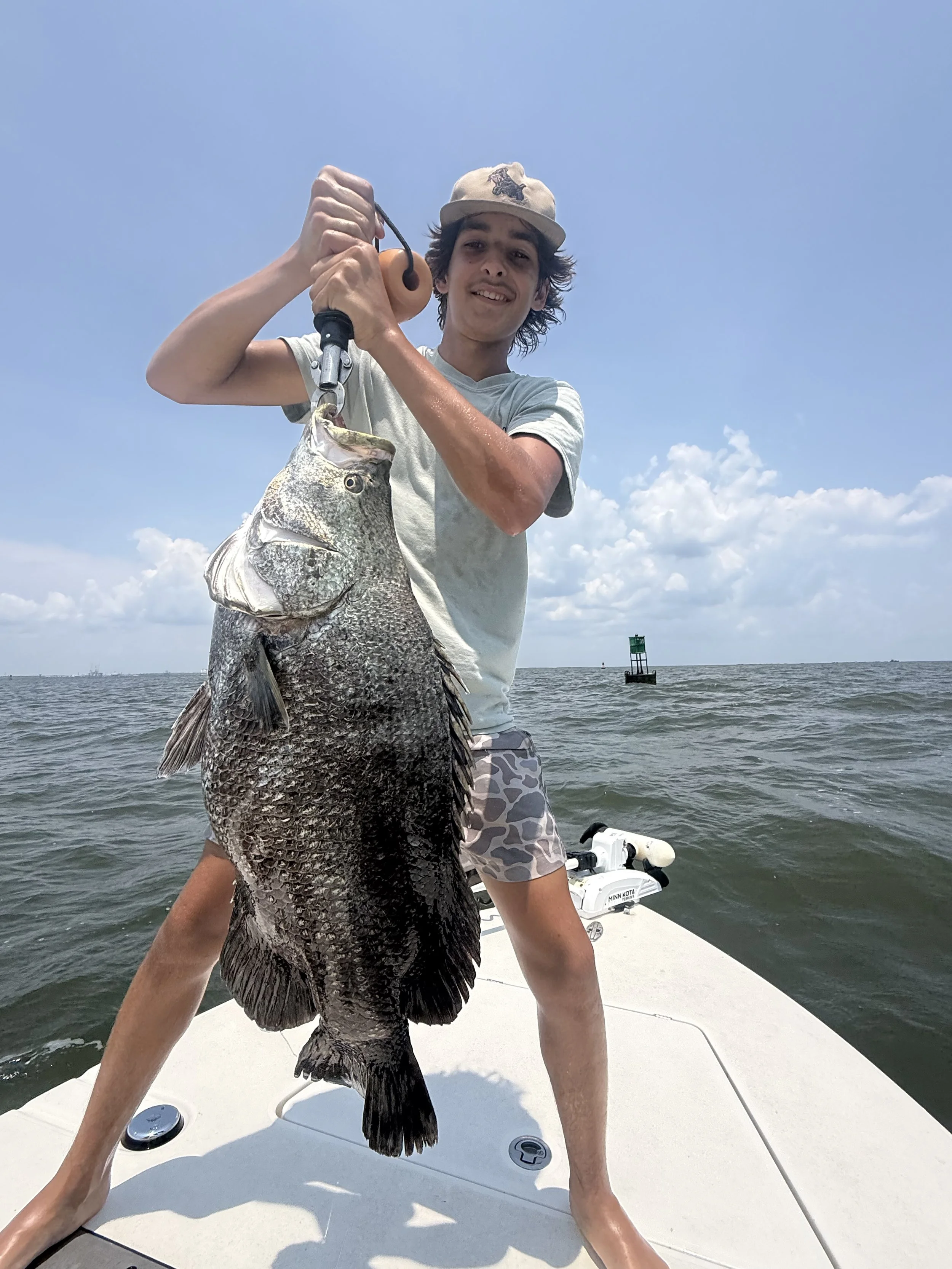 A young man wearing a beige cap and camouflage shorts on a boat holding a large fish with an underbite, while the ocean and sky with some clouds are in the background.