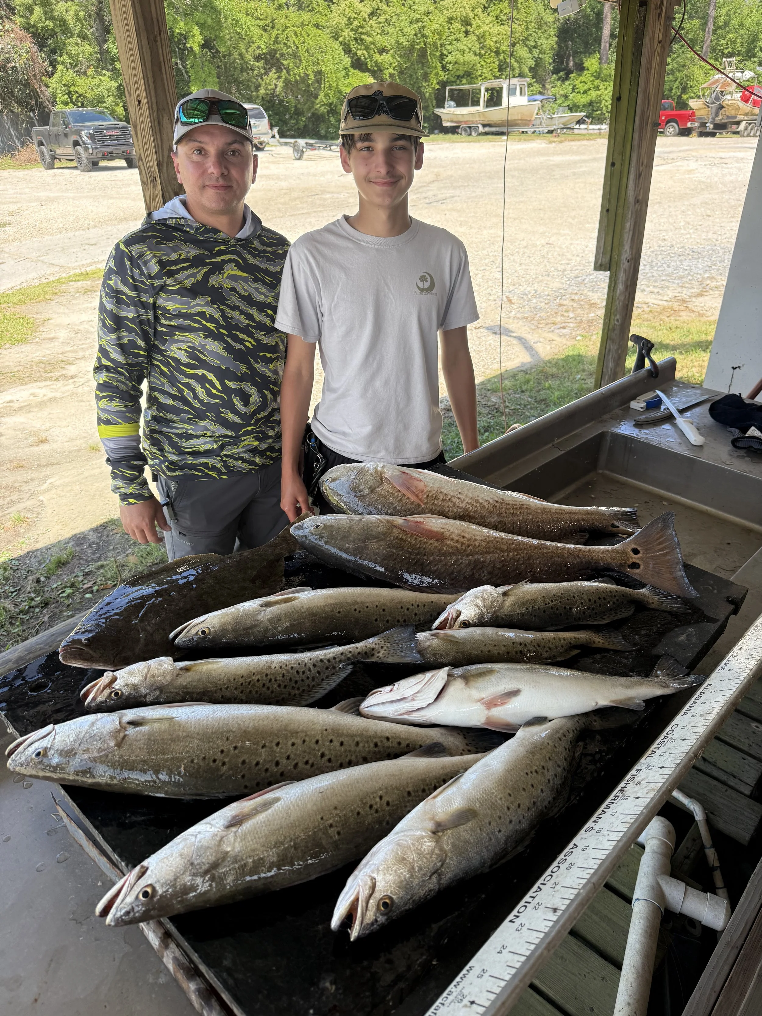 Man and teenage boy standing behind a table with multiple caught fish, outdoors under a wooden shelter with boats and vehicles in the background.