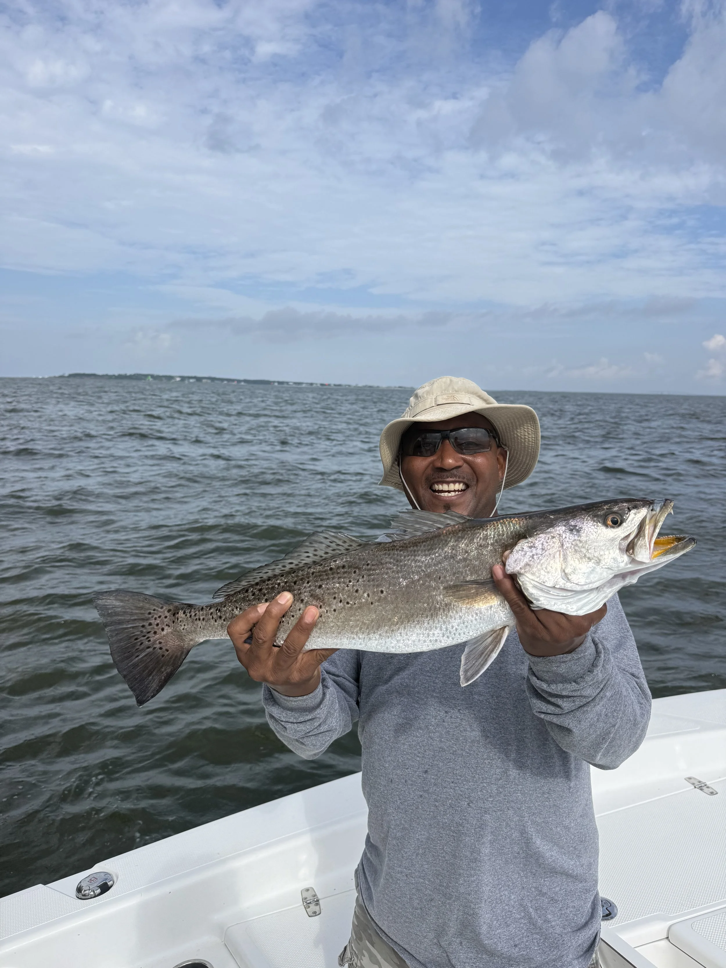 A man wearing a fishing hat and sunglasses, smiling, holding a large fish on a boat with water and a cloudy sky in the background.