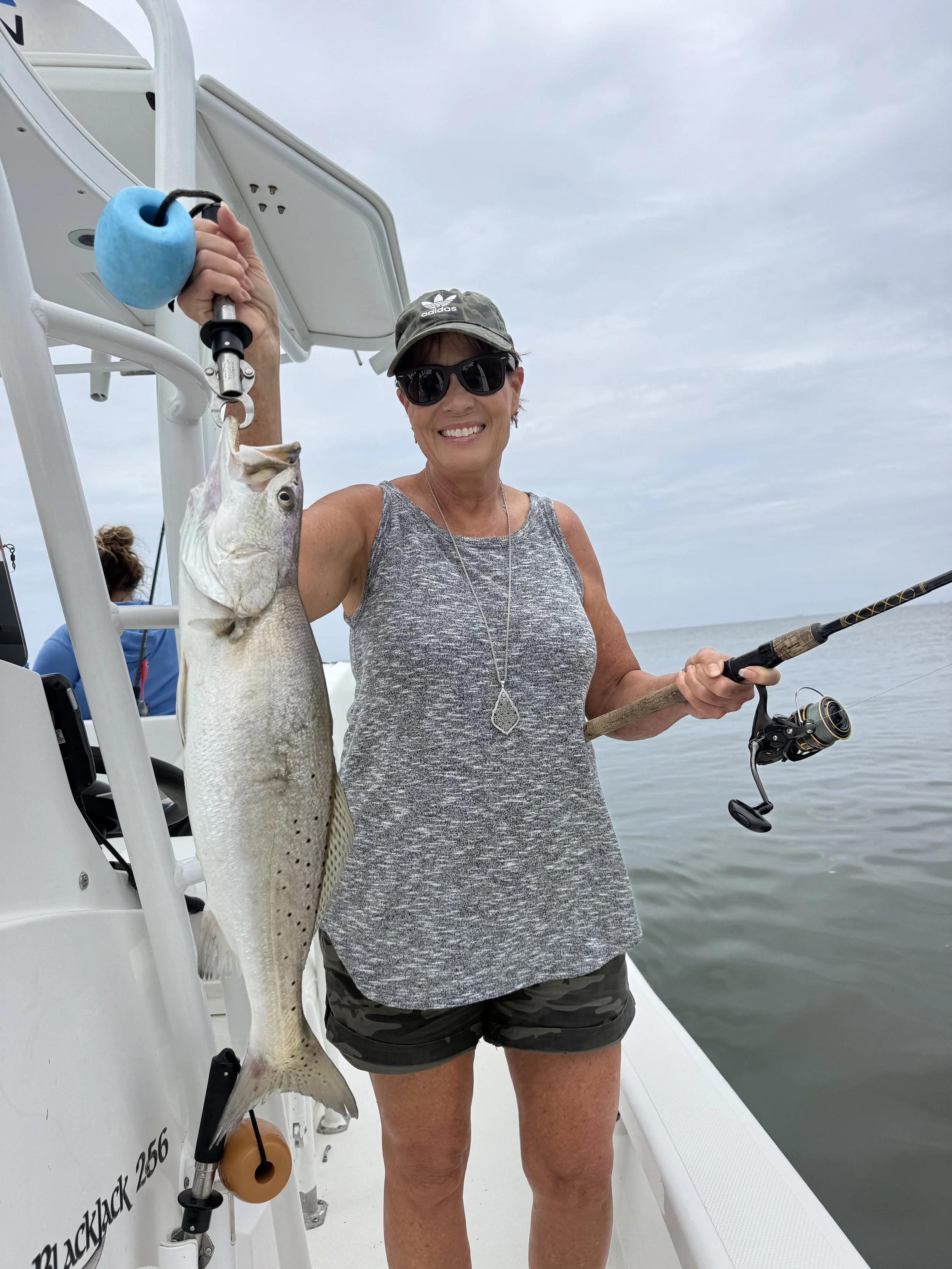 A woman on a boat holding a large fish she caught, wearing sunglasses, a gray tank top, and a camouflage cap, with a fishing rod in her right hand, and the ocean and cloudy sky in the background.