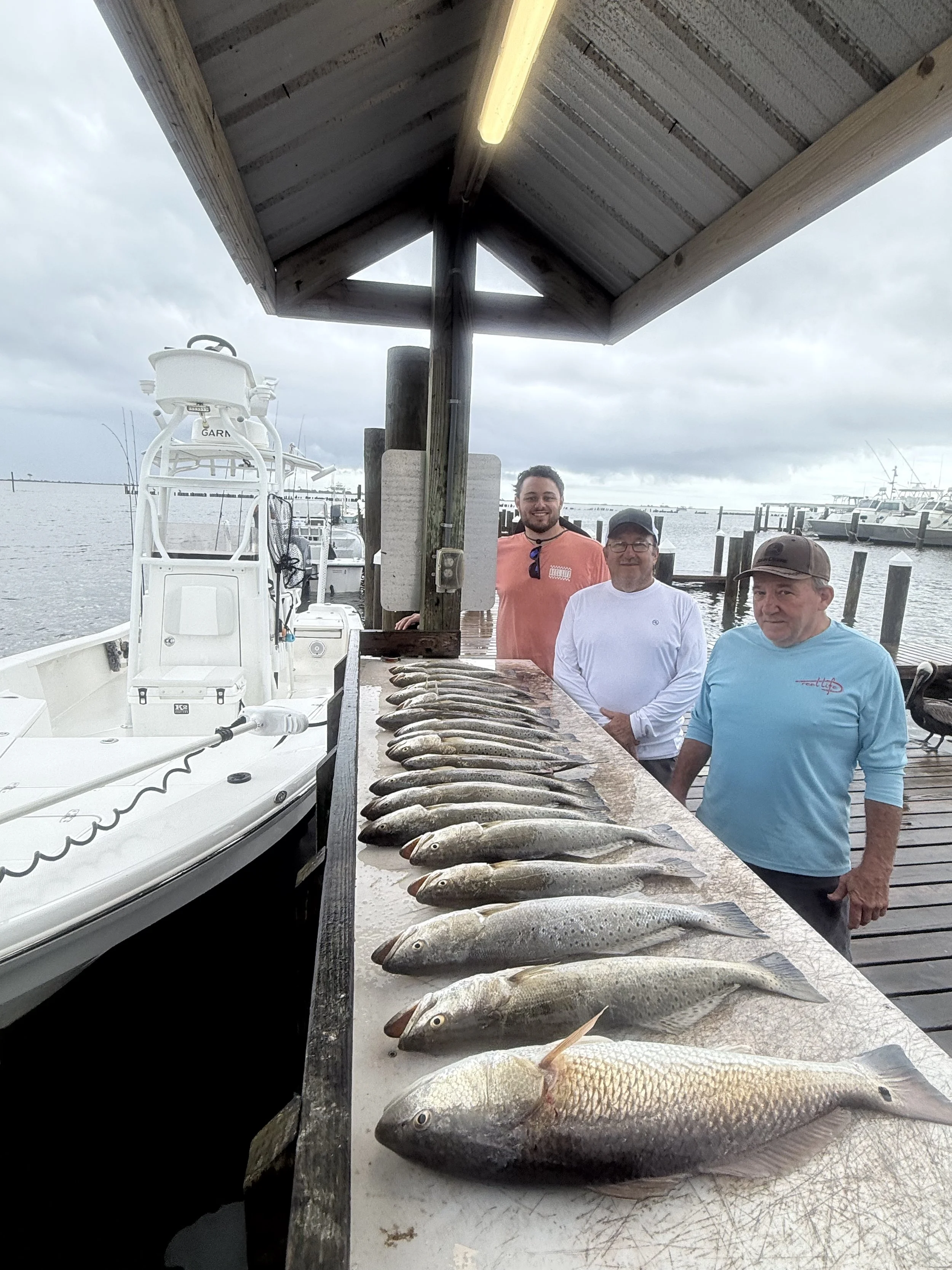 Several fish laid out on a table at a dock, with three people posing behind the fish, a boat and water in the background, under a covered structure.