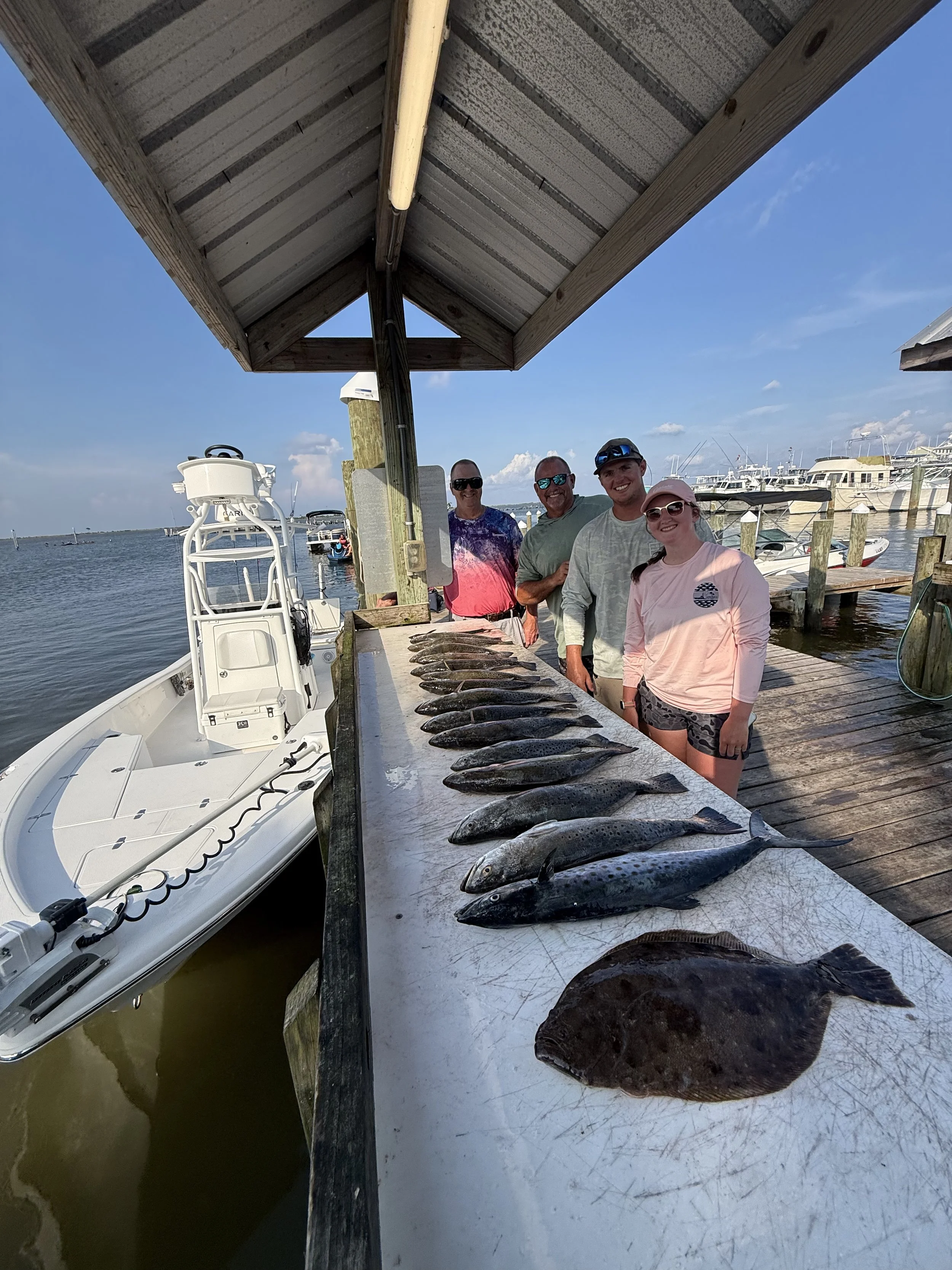 People standing behind a row of fish on a dock with boats and water in the background.