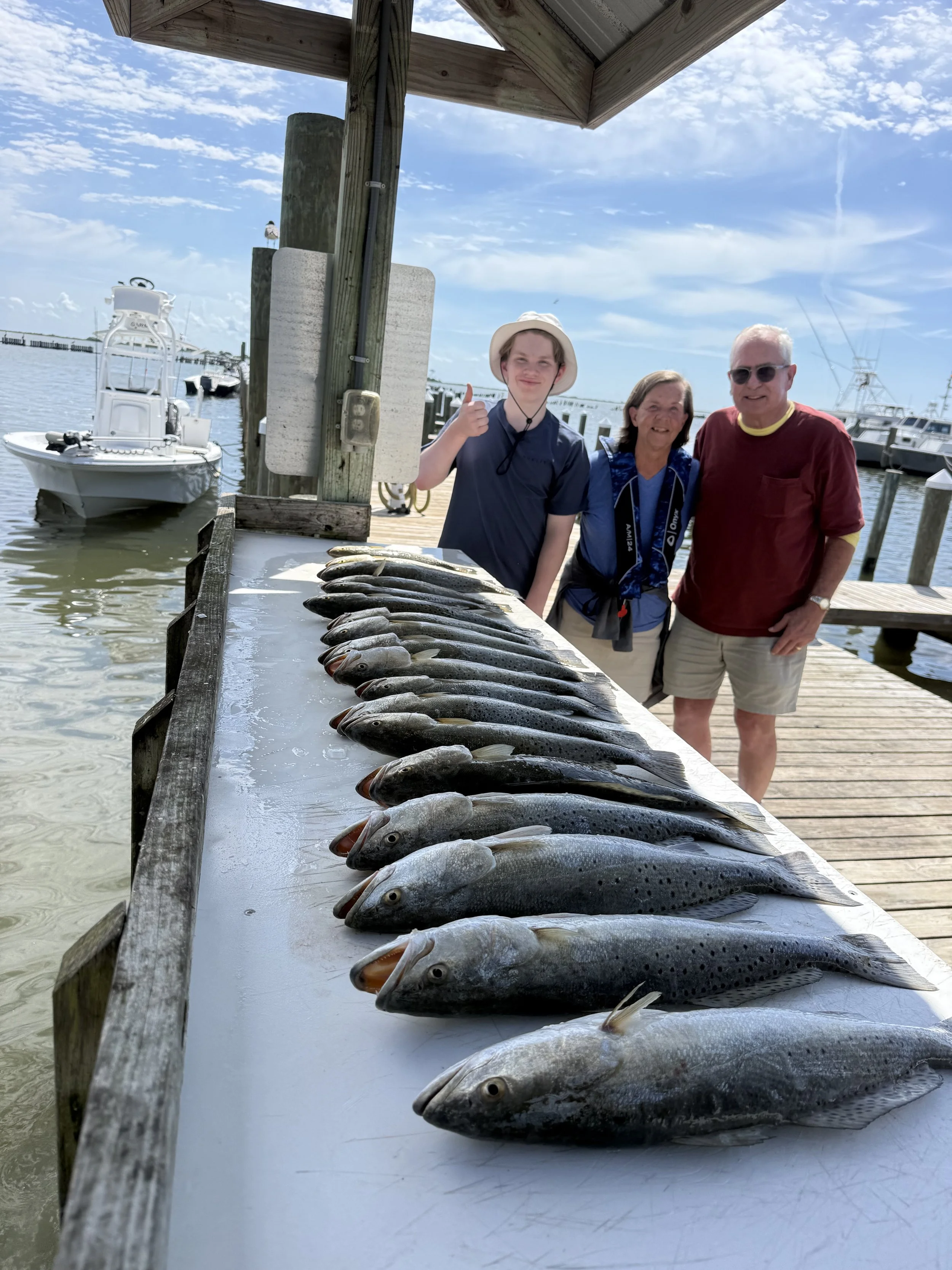 A fish cleaning table with several caught fish laid out on it, near a dock with boats in the background, under a partly cloudy sky.