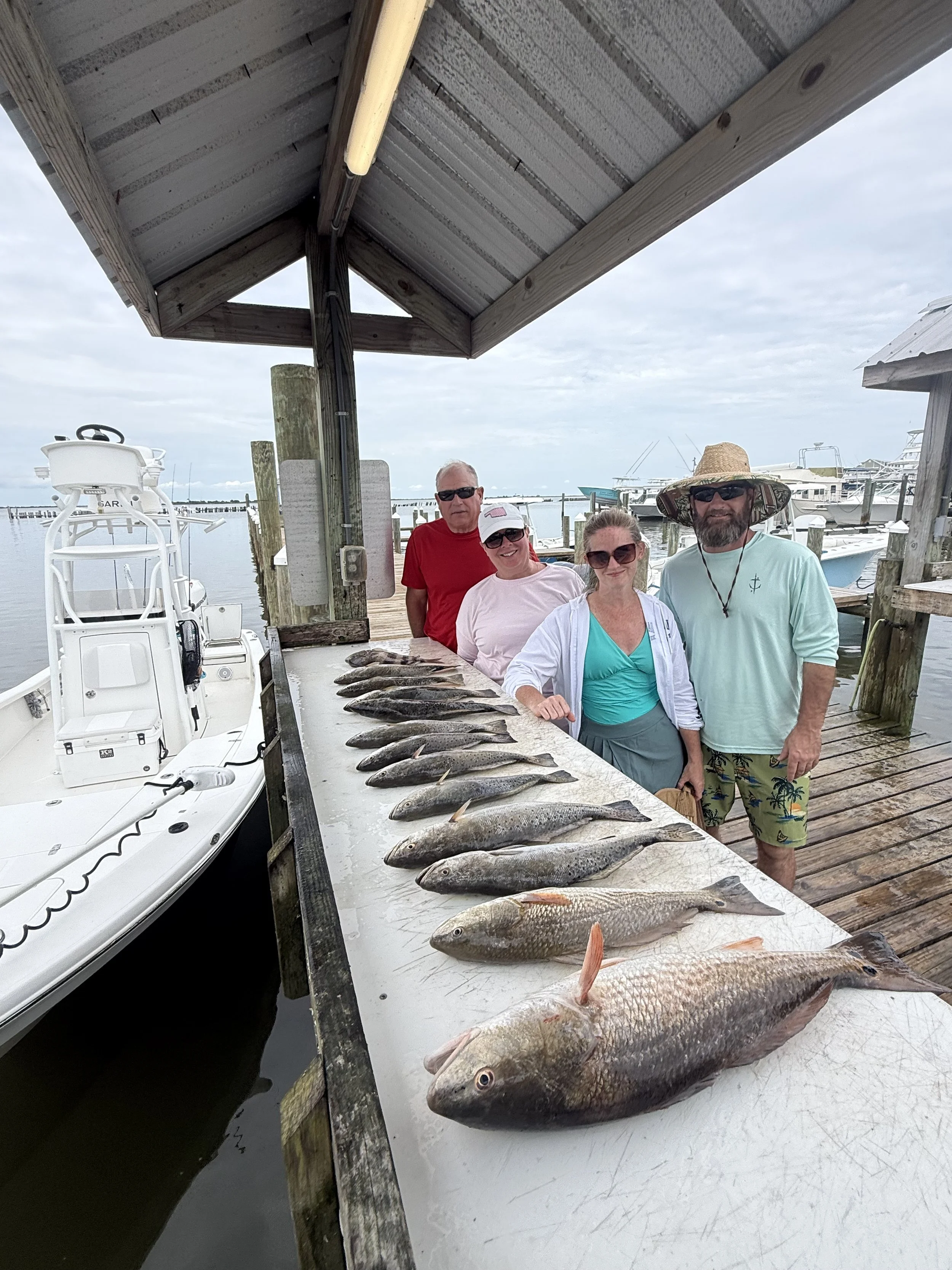 Four people standing behind a large table of freshly caught fish, at a marina with boats in the background. Two men and two women are wearing casual summer clothing and sunglasses.