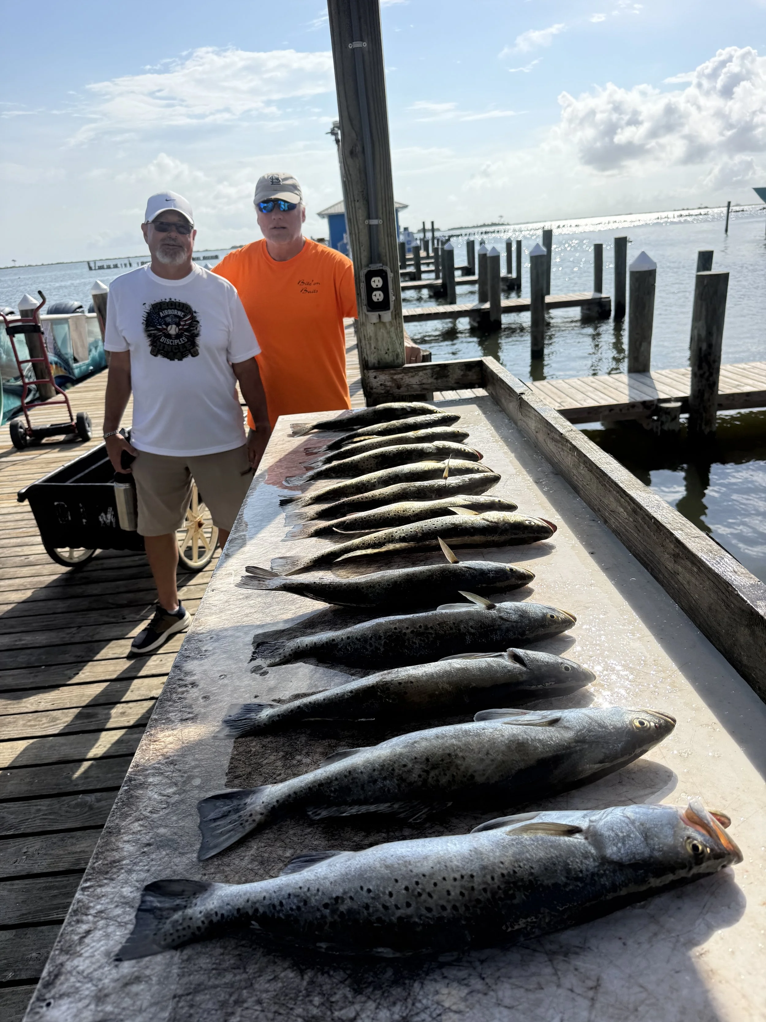 Two men standing behind a table with freshly caught fish on it, on a dock by the water, with a sky and clouds in the background.
