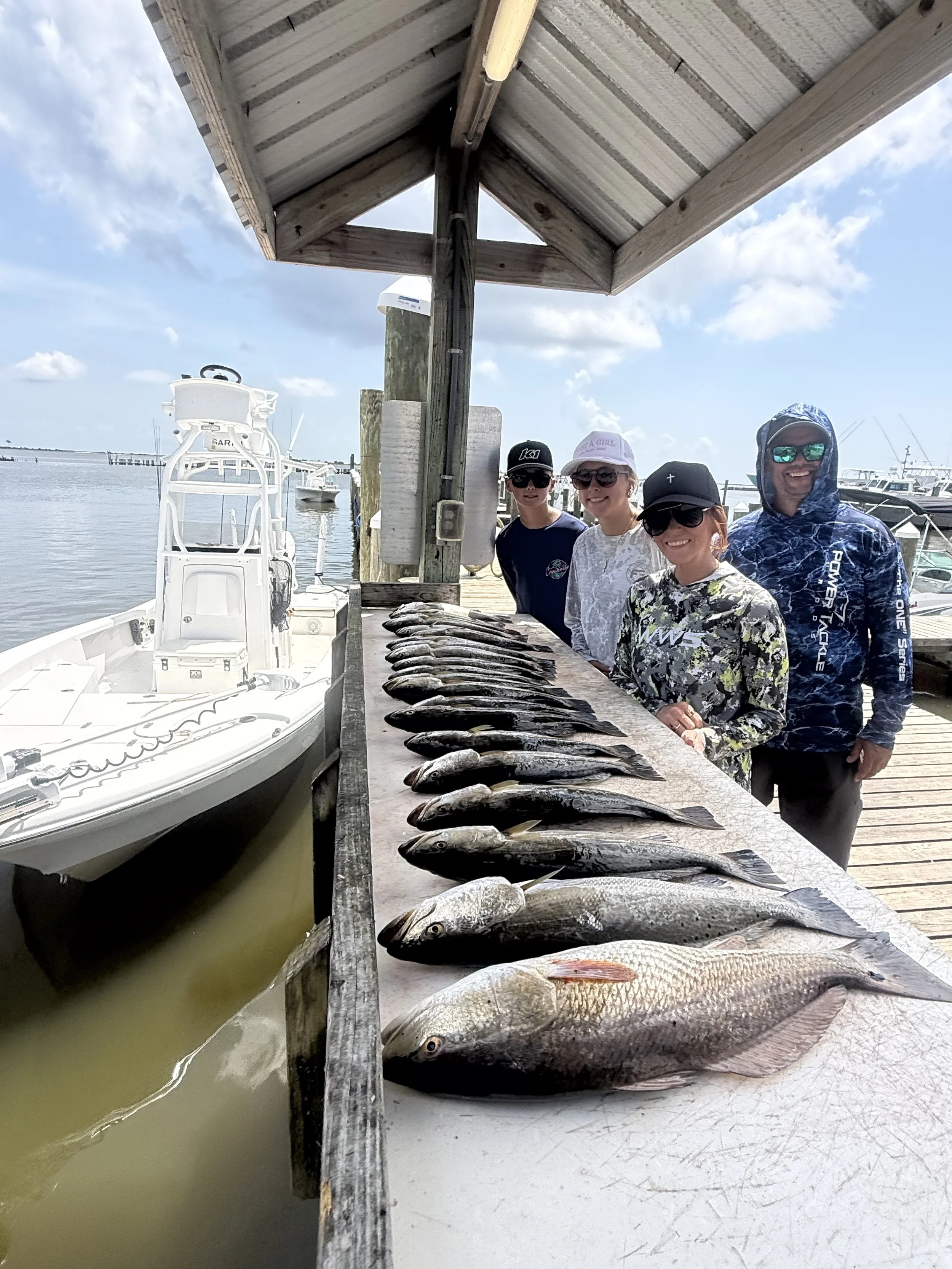 Four people standing behind a row of fish laid out on a dock, with boats and water in the background, under a wooden shelter.