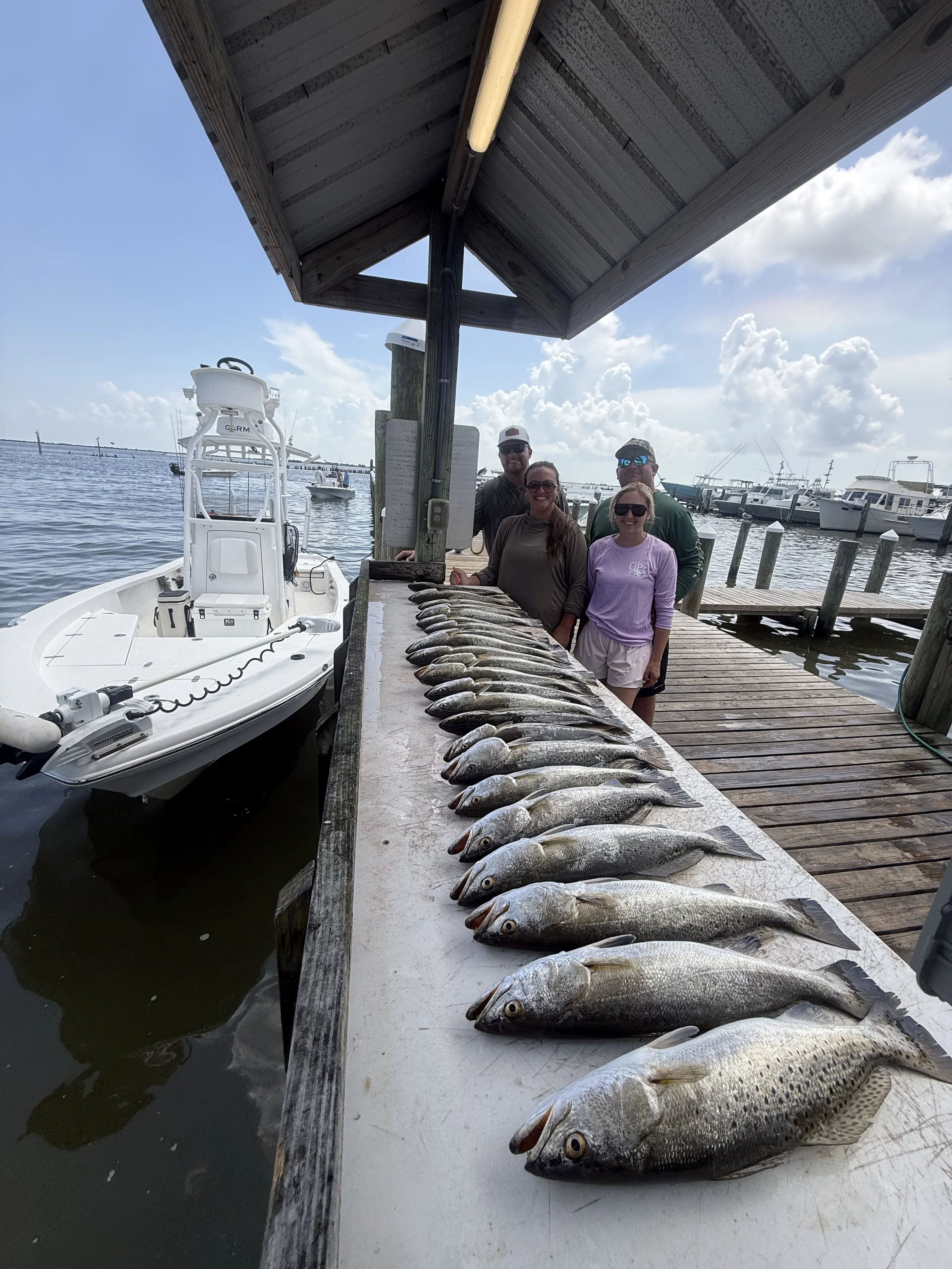 A group of four people standing behind a row of caught fish on a dock by the water, with boats and a cloudy sky in the background.