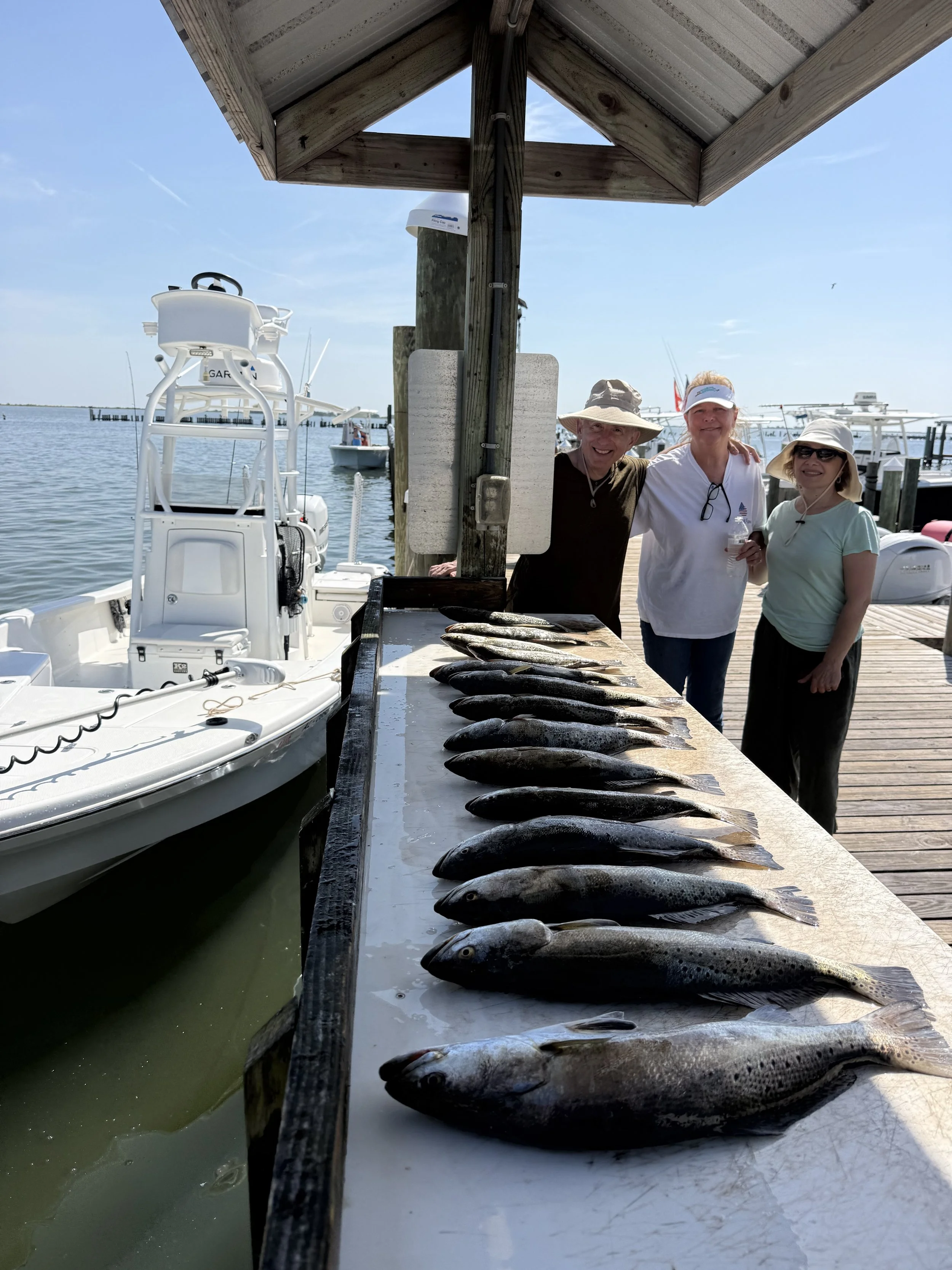 Three people standing next to a table of fish at a dockside with boats in the water and clear sky in the background.