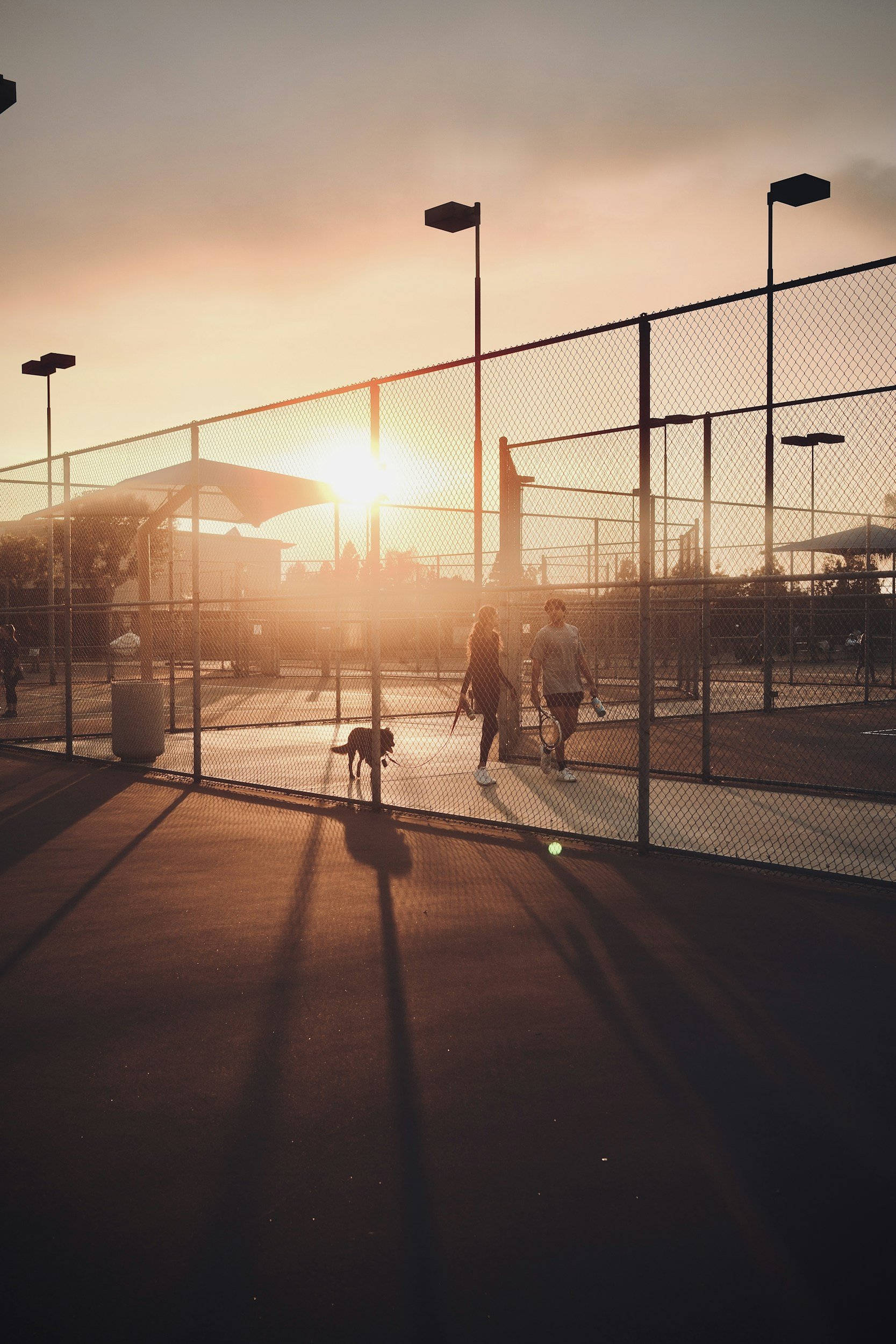 Deux personnesvsur un terrain de tennis en plein air au coucher du soleil, entouré d'une clôture en maille métallique.