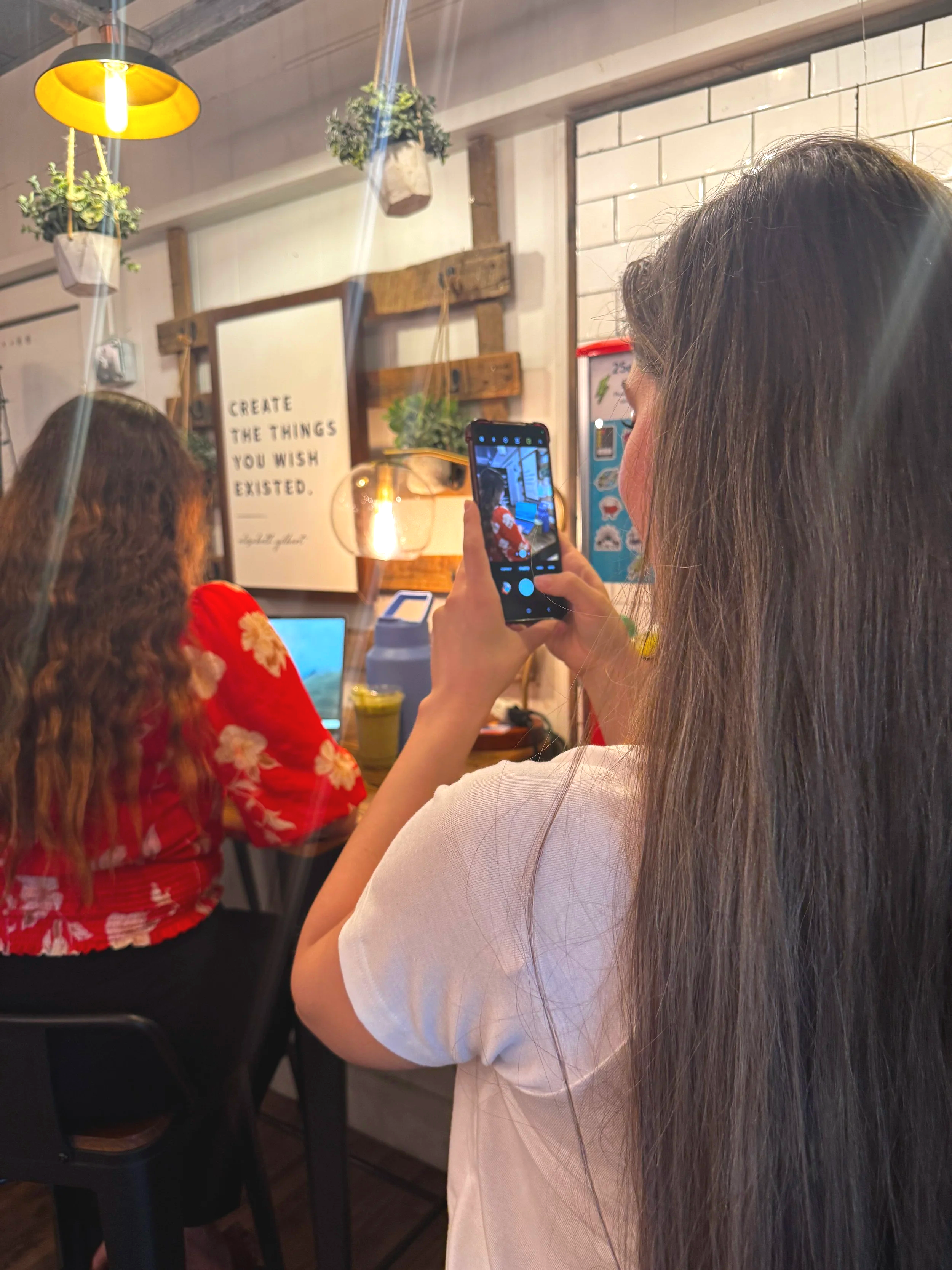 local marketing agency team member taking a photo with her smartphone inside a cafe, where a person in a red floral shirt is sitting at a table, and there are decorative hanging plants.