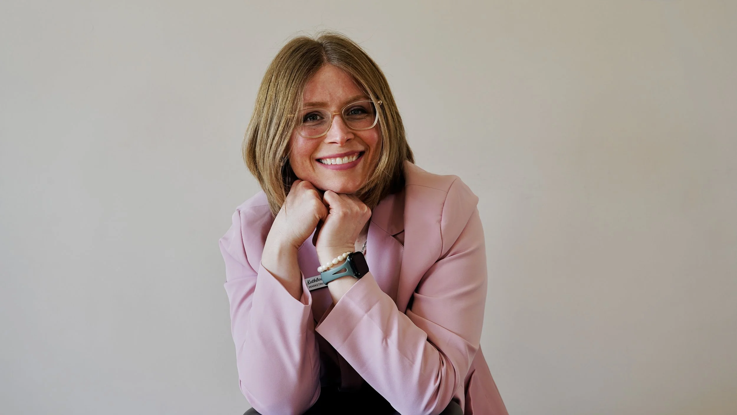 A woman with shoulder-length light brown hair, wearing glasses and a pink blazer, smiling, with her chin resting on her hands, seated in front of a plain light-colored background.