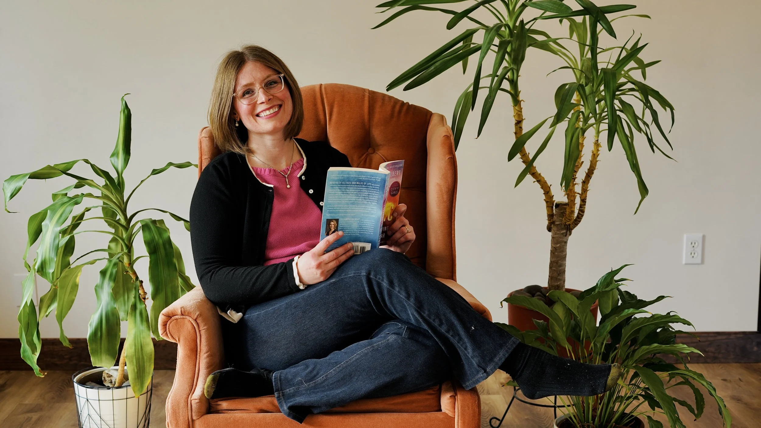 MadeUp Media founder Kathleen Haney smiling in an orange armchair, reading a book among green houseplants