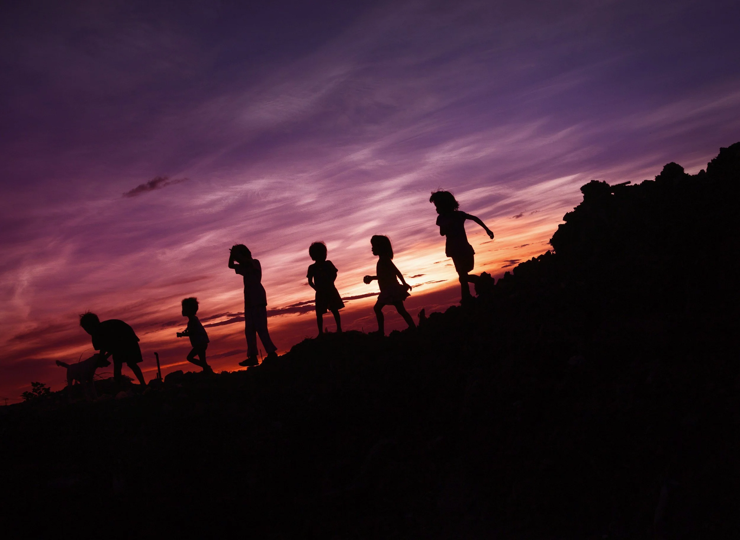 Silhouettes of children walking on a hillside during sunset with colorful pink, purple, and orange sky.