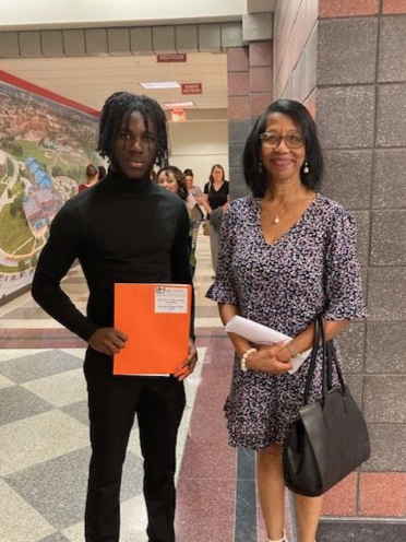 A young man and an older woman standing together in a hallway, smiling at the camera. The young man is holding an orange folder, and the woman is holding papers and a black purse.