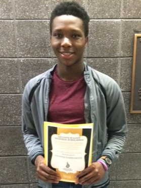 Young person smiling, holding a yellow certificate or diploma, standing against a brick wall.