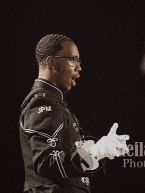 Side profile of a young man wearing glasses and a drum major uniform, with white gloves against a black background.