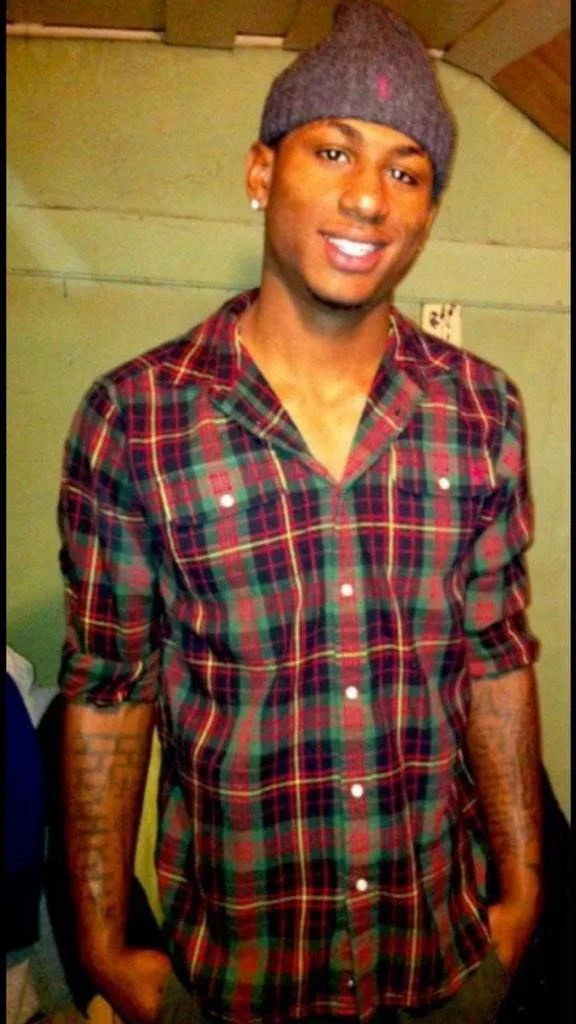 Young man smiling, wearing a gray beanie and a red and green plaid shirt, standing indoors against a green wall.