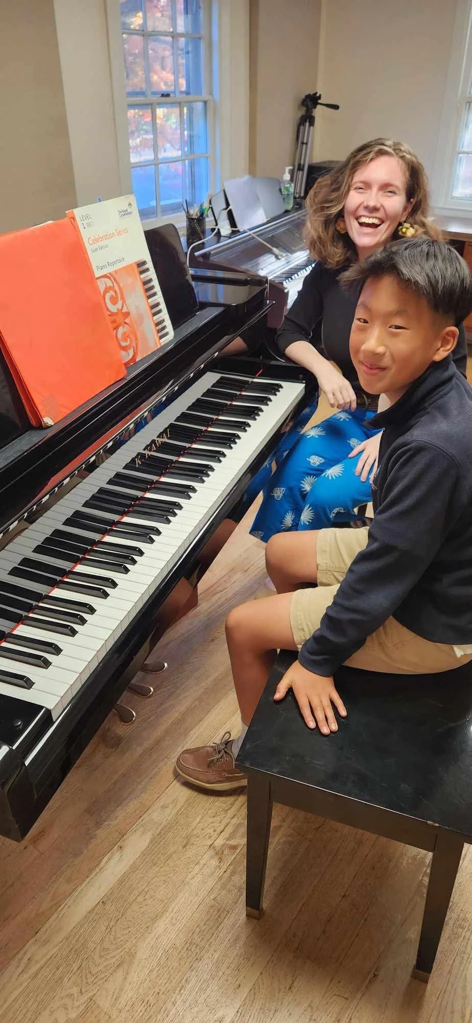 A woman and a young boy sitting at a piano in a room with large windows. The woman is smiling widely and the boy is sitting on a bench, looking sideways with a slight smile. There are music books on the piano, and a tripod with a camera is visible in the background.