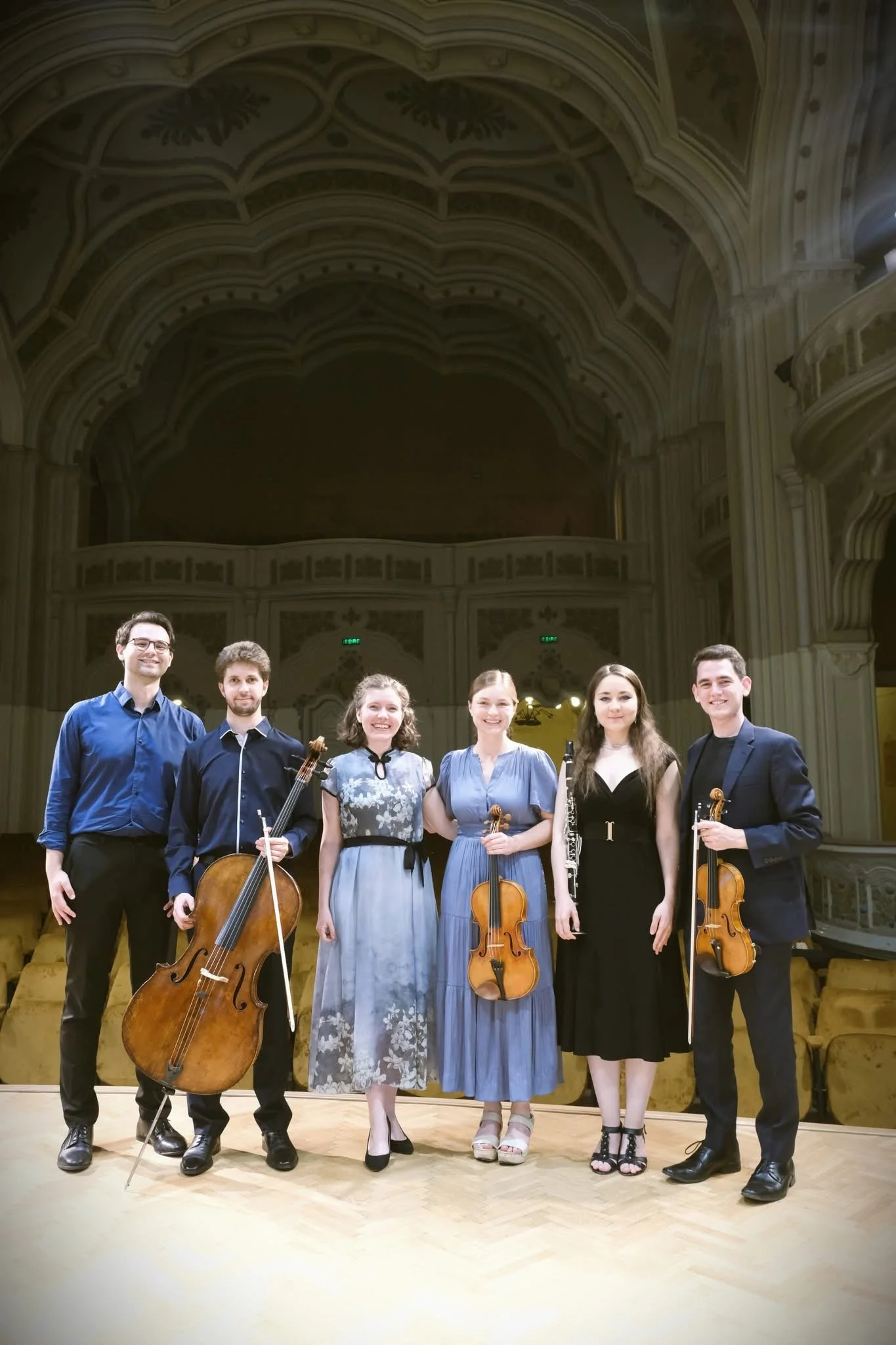 Six musicians holding string instruments on stage in an ornate concert hall.