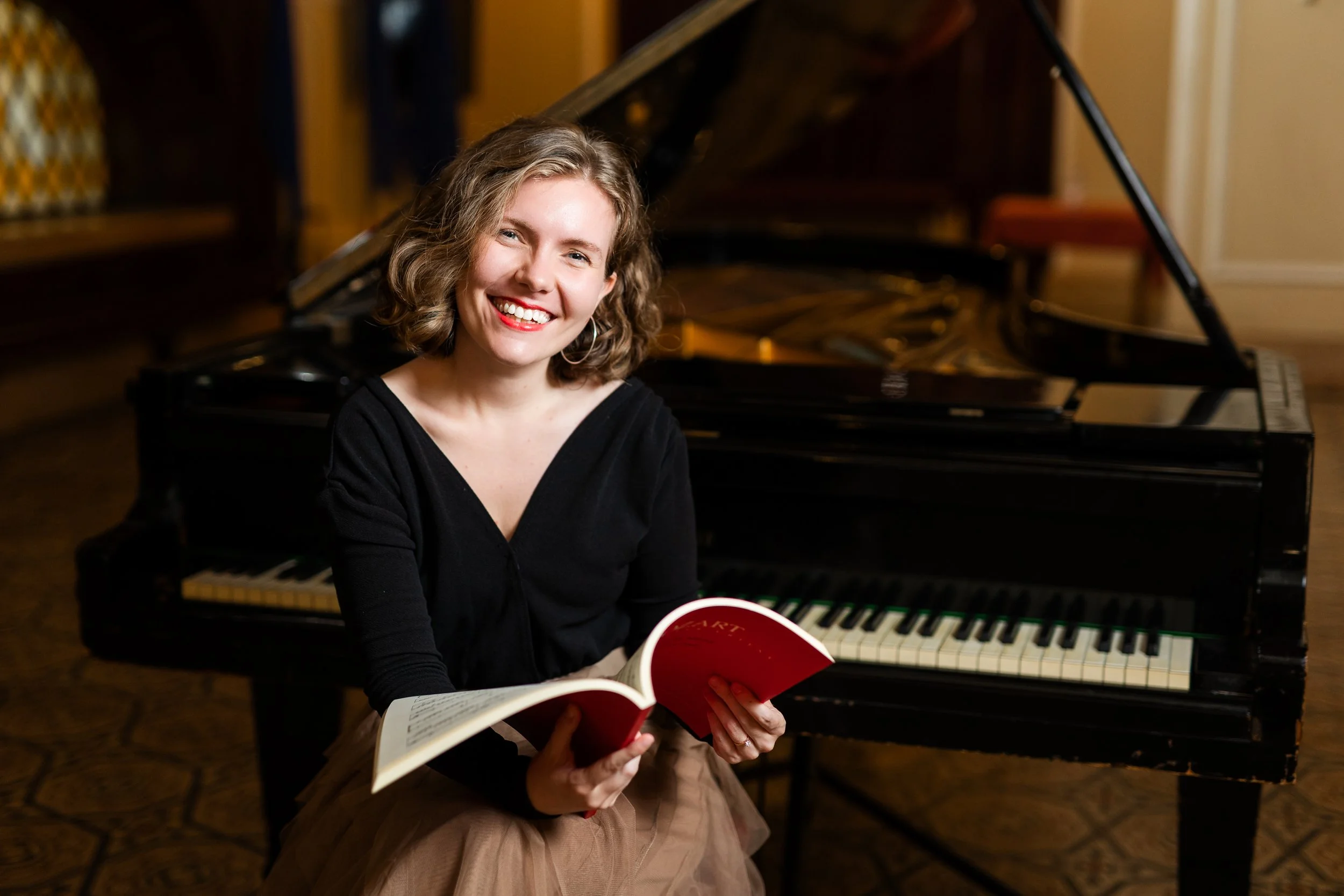 Young woman with curly hair smiling while sitting in front of a grand piano, holding an open music book.