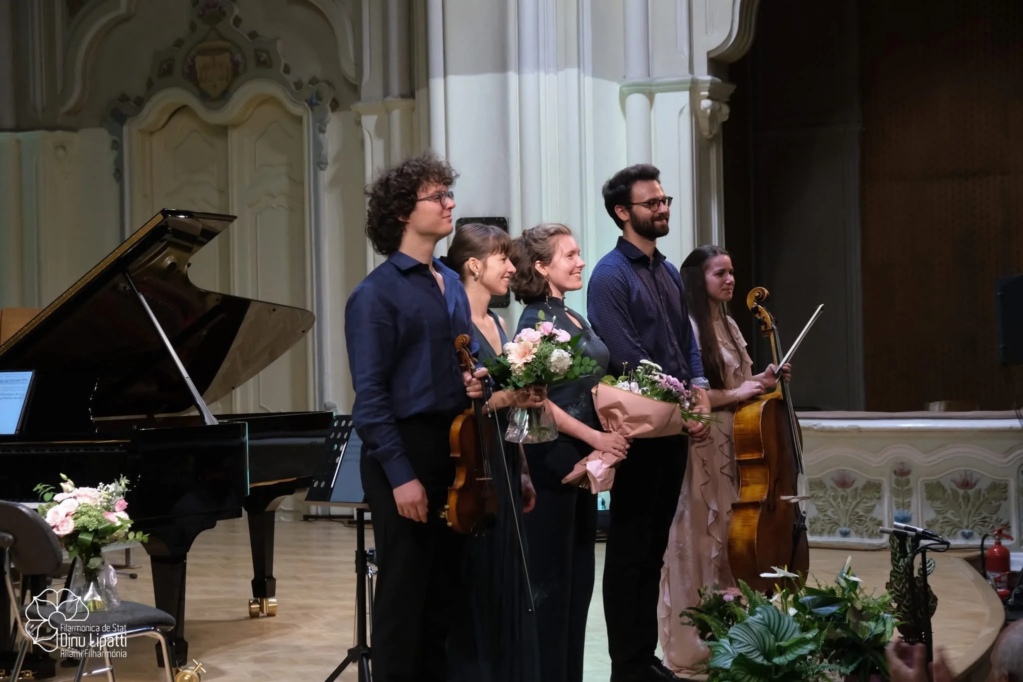 Six musicians standing on stage with flowers, a grand piano, and musical stands in a concert hall.