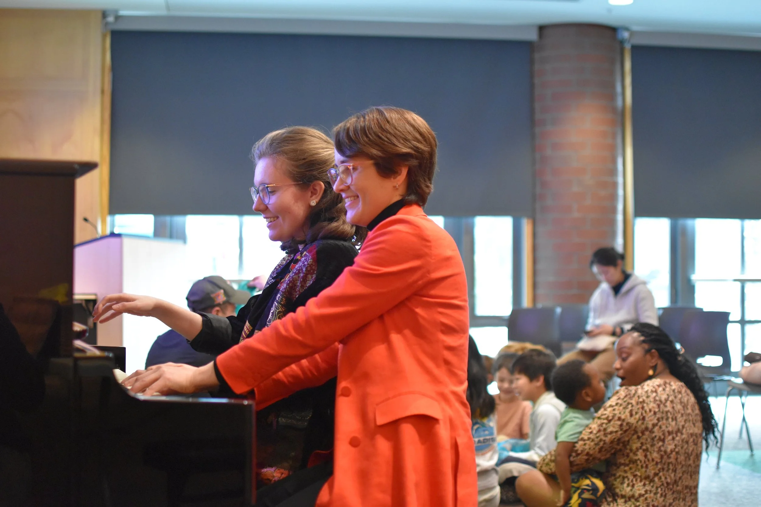 Two women playing piano together in a lively indoor setting with children and adults in the background, and large windows letting in natural light.
